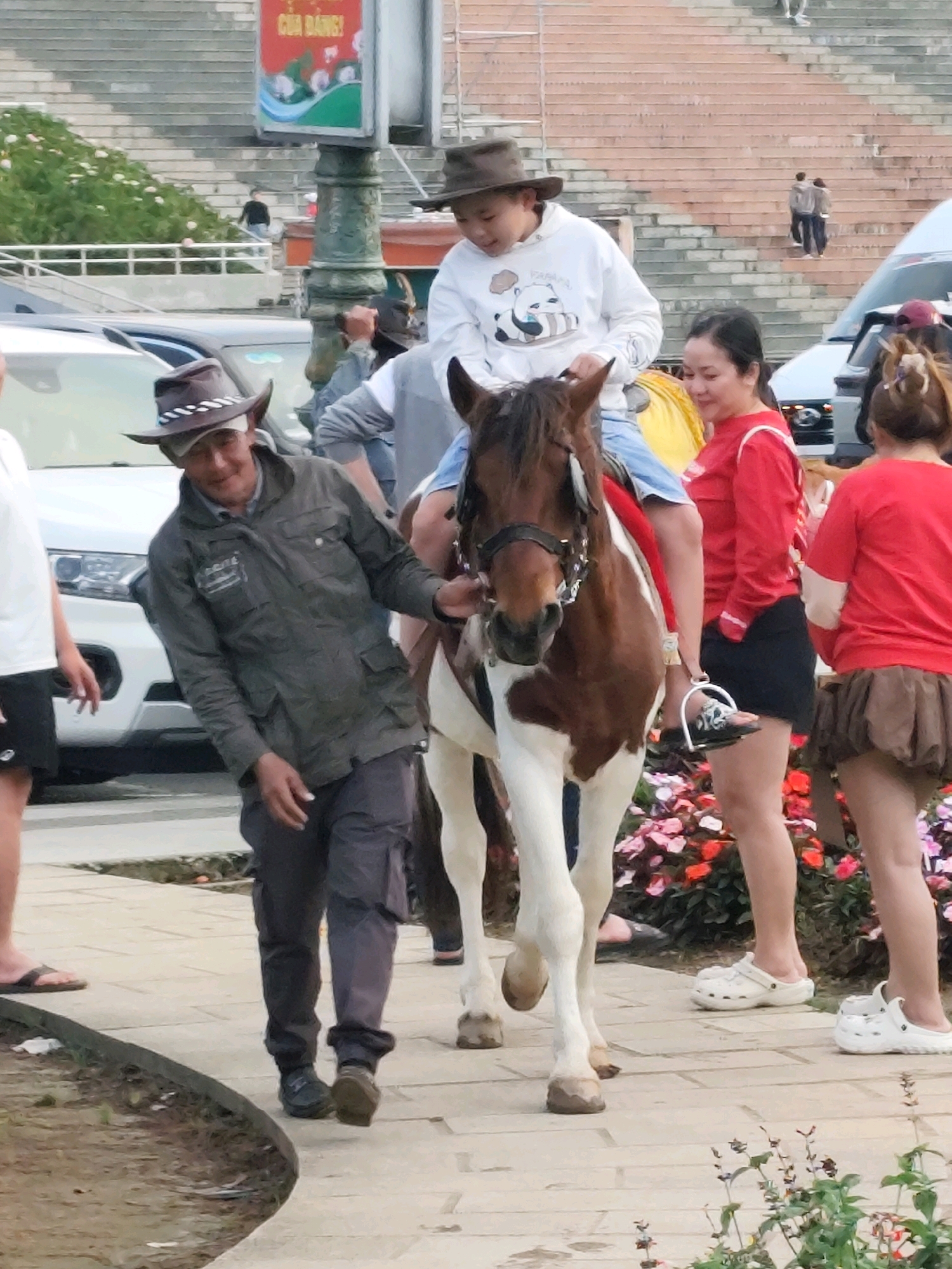 In Da Lat, Vietnam, near a stone staircase and a traffic light, a piebald horse is being led by a man wearing a brown cowboy hat and a green jacket. A child in a white hooded sweatshirt with a panda graphic is riding the horse. To the right, two women in red tops and dark bottoms are standing, and a third woman in a red top and a brown skirt is further to the right. The ground is paved with light-colored stones, and a border of colorful flowers is visible. A white car is parked to the left of the scene.