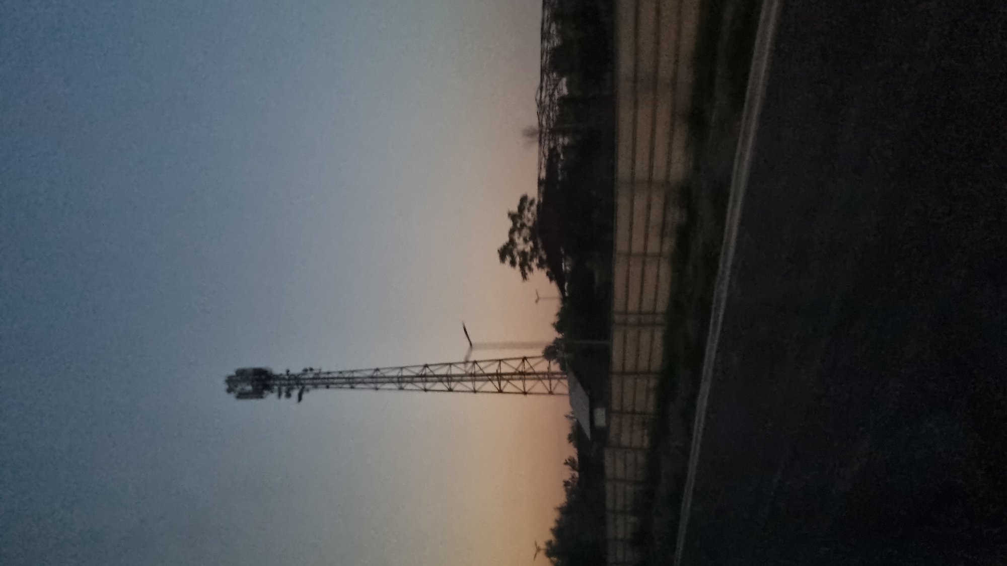 This image shows a communication tower against a twilight sky. The tower is tall and lattice-like, with antennae at the top. To the left of the tower is a small building. In the background, there are trees and a large, partially constructed structure that appears to be a building or stadium. A concrete fence runs horizontally across the middle ground. In the foreground, a dark asphalt road is visible, suggesting the image was taken from a moving vehicle. The overall impression is of an industrial or semi-developed area on the outskirts of a city, possibly at dusk.