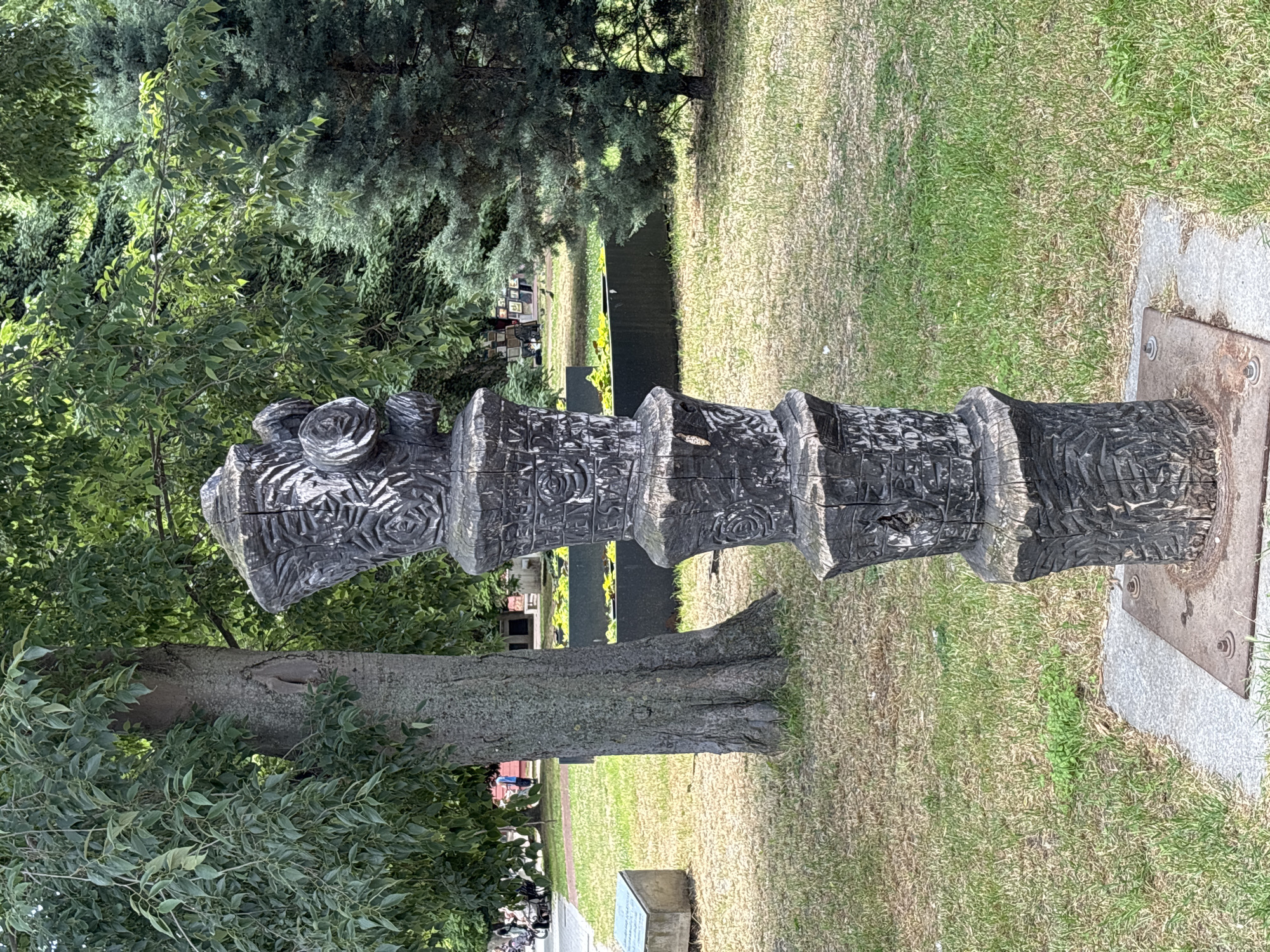 The image shows a tall, carved wooden sculpture situated in a grassy area. The sculpture is dark brown, weathered, and has intricate patterns carved into its surface, including floral motifs. It stands on a concrete base secured by metal plates and bolts. Behind the sculpture, a large tree with textured bark is visible, along with lush green foliage. Further back, there is a dark rectangular planter with yellow water lilies, a path, and some people in the distance. To the right of the sculpture, evergreen trees form a dense background. The environment appears to be a park or public garden in the city of Varna, Bulgaria.
