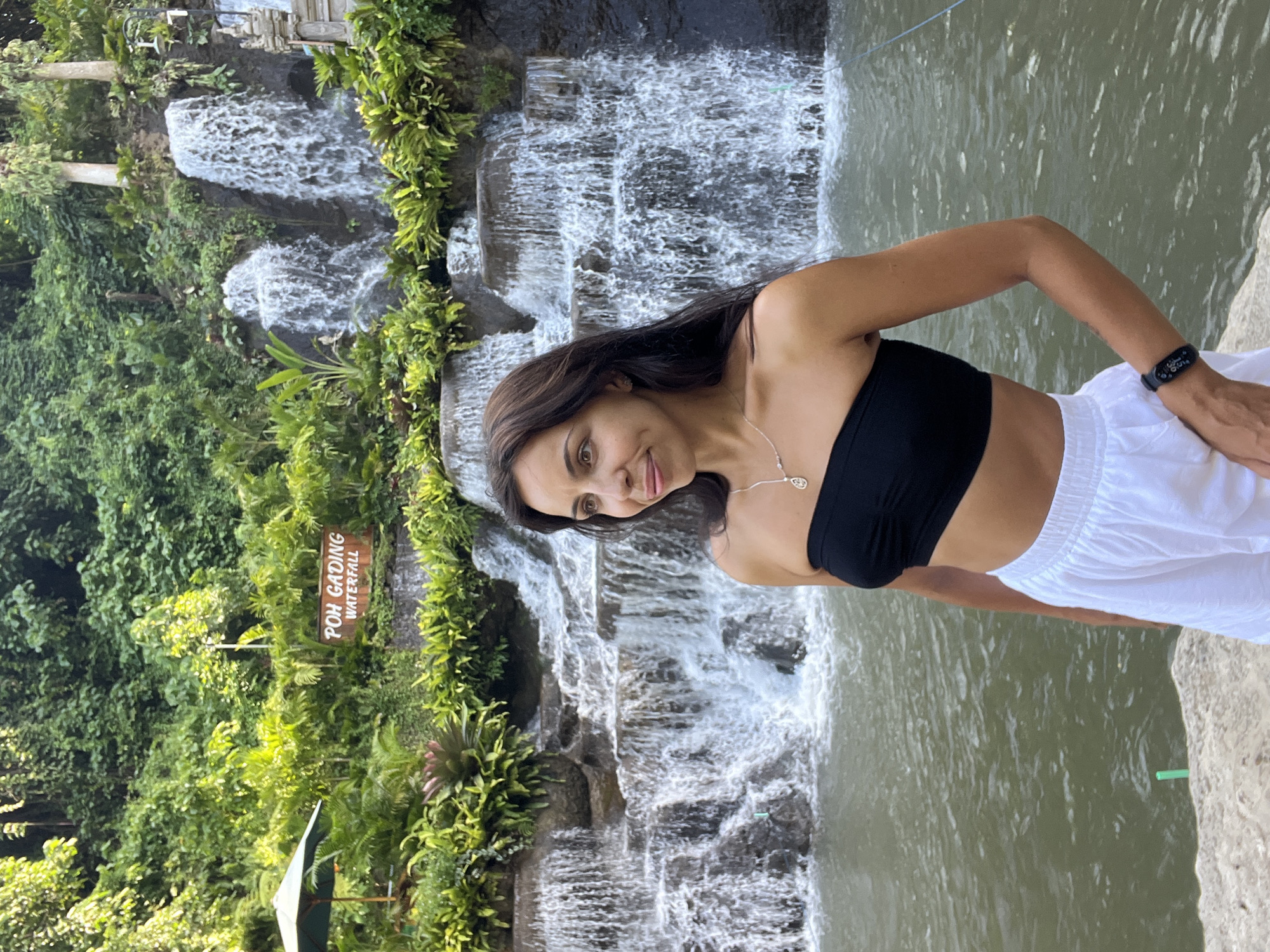 A medium shot shows a woman in a black top and white skirt standing in front of a waterfall. The waterfall is multi-tiered, and a body of water is present on the right side of the waterfall. Vegetation and a sign that reads "POH GADING WATERFALL" are visible on the left side of the waterfall. The image was taken in Sukawati, Indonesia.