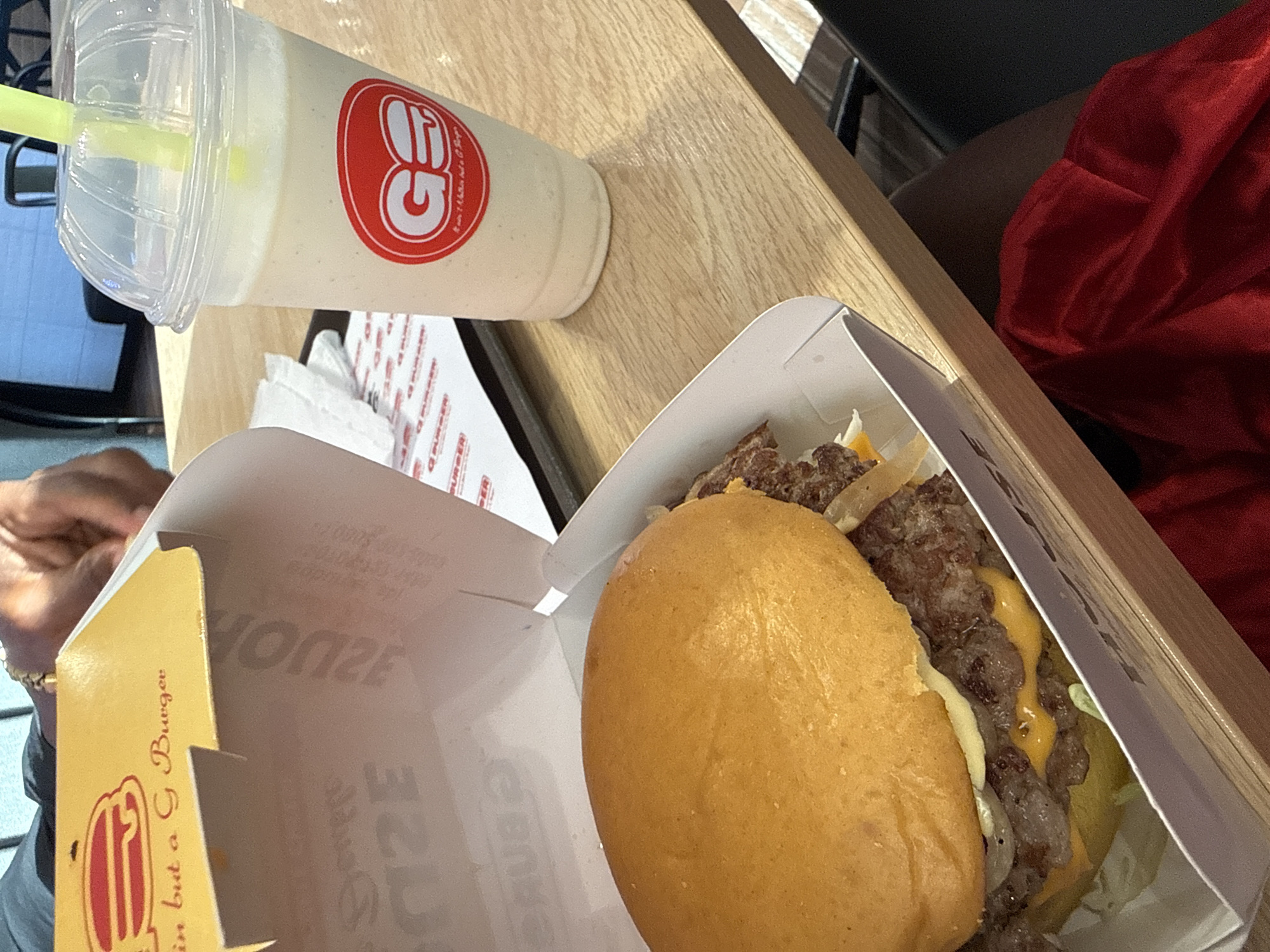 The image depicts a close-up of food items from a restaurant called "GBurger," likely in Abuja, Nigeria, based on the brand name on the packaging. The scene includes a burger in a white cardboard box, a milkshake in a clear plastic cup with a yellow straw, and a stack of branded paper napkins. The burger appears to have a beef patty, cheese, lettuce, and onions on a yellow bun. A portion of a person's hand and arm is visible on the left side of the frame, with a gold-colored bracelet on the wrist. Another person's legs are visible in the background. The food and packaging are placed on a wooden table. The overall setting suggests a casual dining environment.