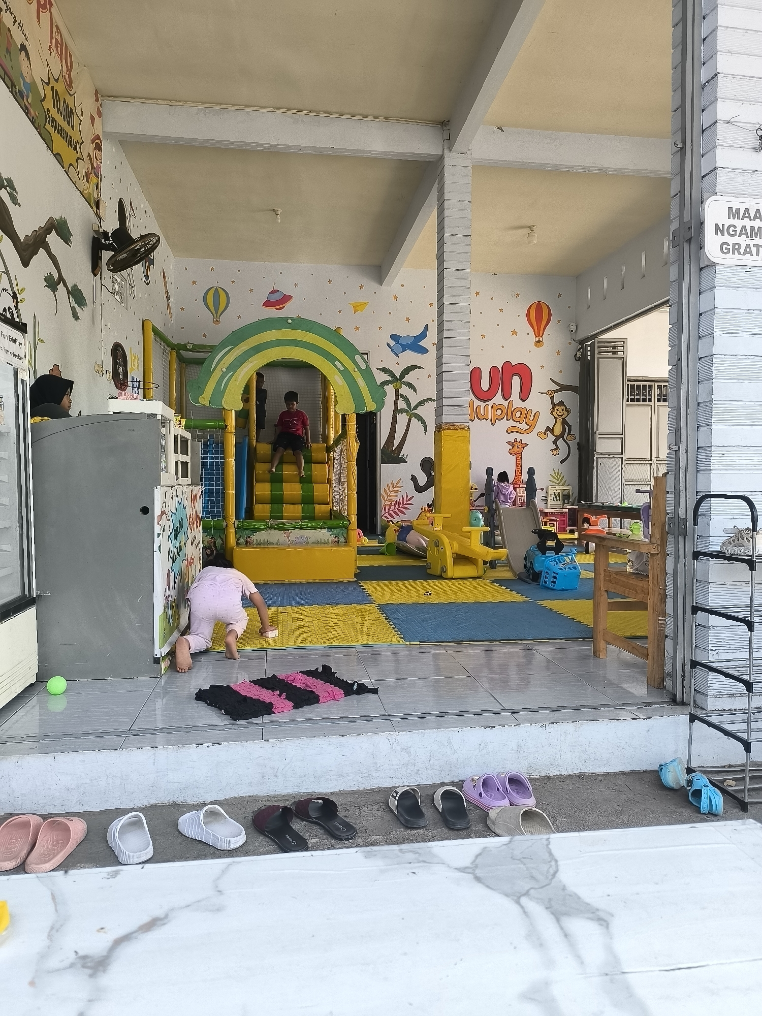 The image depicts a brightly colored indoor playground at Fun Eduplay, located in Dagangan, Indonesia. The playground features a yellow and green structure with stairs, a slide, and a ball pit. Several children are visible: one is on the stairs, another is playing on the floor, and a third is partially visible near a cabinet. The walls are decorated with cartoon murals of animals, hot air balloons, and airplanes. In the foreground, a row of shoes and sandals is arranged outside the playground entrance. The overall scene conveys a lively and playful atmosphere suitable for young children.