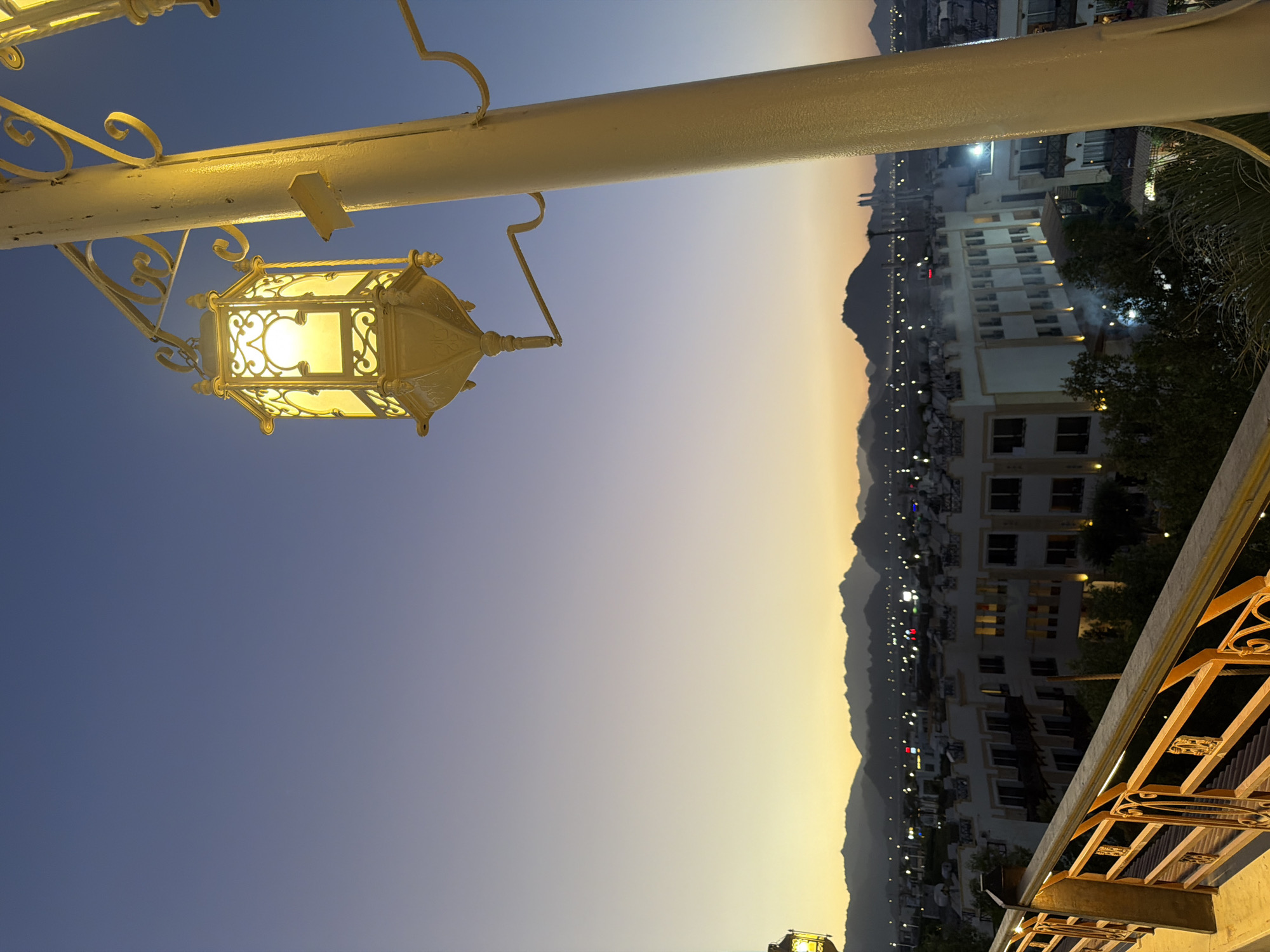 A lamppost with an ornate, hexagonal lantern in the foreground is lit, casting a warm yellow glow against a clear blue sky transitioning to a pale orange horizon. The lantern is suspended by a decorative metal arm attached to a thick, cream-colored pole. In the middle ground, a town or city skyline is visible at dusk, with buildings that appear to be hotels or residential complexes, characterized by white facades and dark-framed windows. Numerous lights are illuminated in the distance, suggesting the early evening. A range of mountains forms the backdrop, silhouetted against the fading light. A railing with decorative metalwork is visible in the lower left corner, suggesting the photo was taken from an elevated vantage point, possibly a balcony or walkway. The overall scene depicts a peaceful evening in Sharm El Sheikh, Egypt.