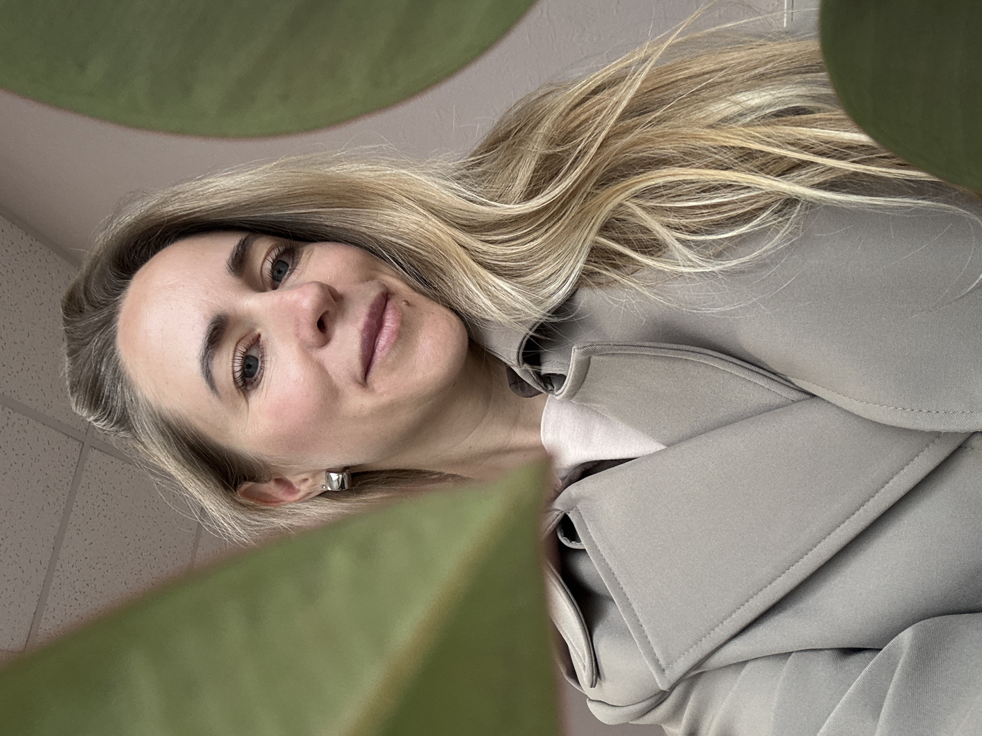 A low-angle, tilted portrait captures a woman with long, wavy blonde and light brown hair looking directly at the camera with a gentle smile. Her blue-grey eyes are open, and she wears light makeup, including mascara and a hint of blush and lip color. She appears to be middle-aged. She is dressed in a light beige or taupe trench coat or jacket over a lighter top, and a small, silver-toned hoop earring is visible in her right ear.
In the immediate foreground, large, blurred green leaves of a plant frame the bottom and left side of the image, suggesting the photo was taken from behind or under the foliage. The background features a light-colored tiled ceiling and what appears to be a light-colored wall or surface, indicating an indoor setting, possibly an office or public building in Chaikovsky, Russia. The lighting is bright, consistent with daytime. No visible text is present in the image.
