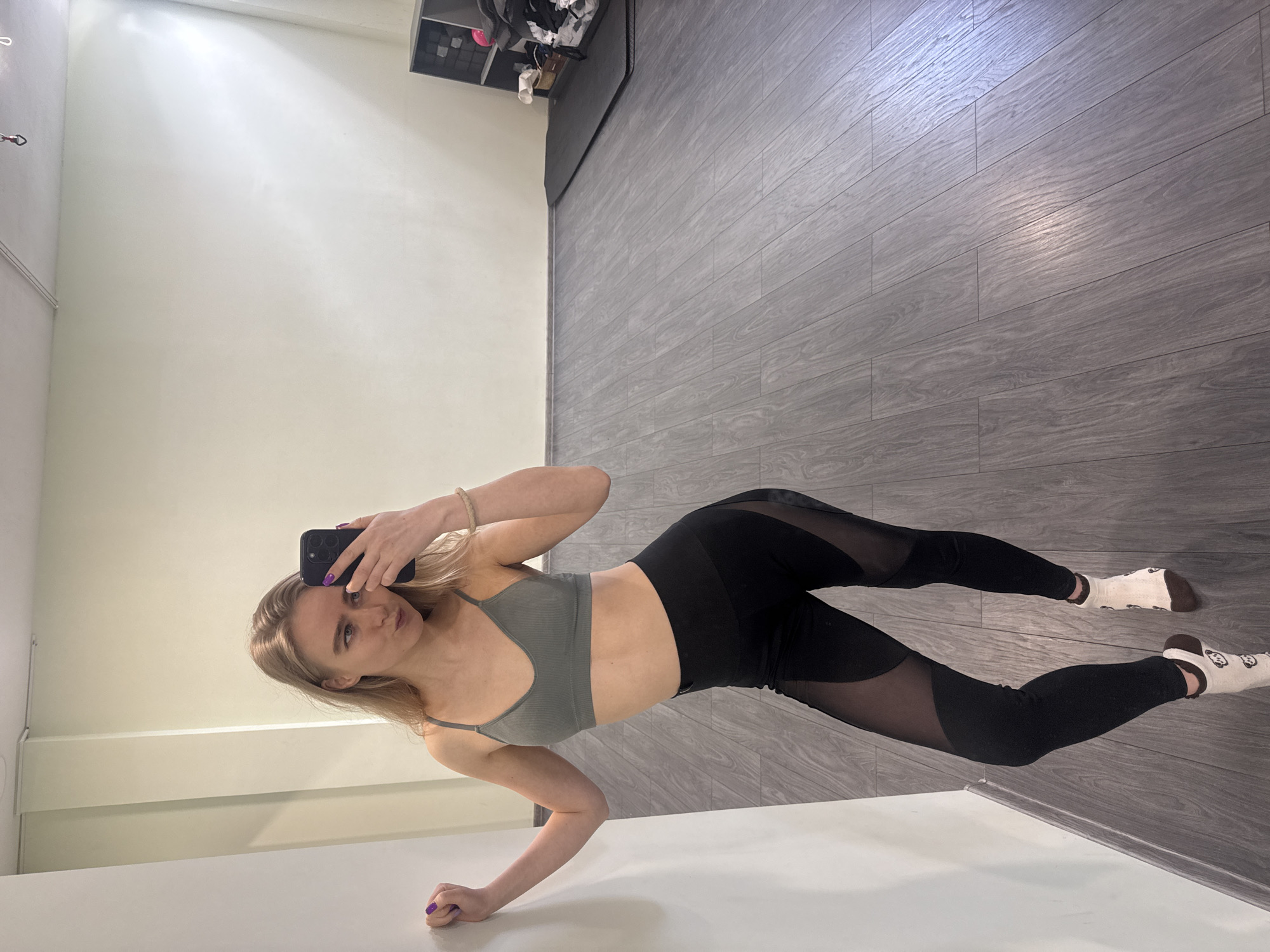 A young woman with blonde hair, wearing a grey ribbed sports bra and black leggings with sheer mesh panels, takes a mirror selfie indoors. She holds a dark-colored smartphone in her right hand, positioned to capture her reflection. Her left arm is bent, resting on a white counter or surface in the foreground. She wears white socks with a brown, possibly animal, pattern and has purple nail polish on her left hand.
The setting appears to be a room with light, off-white walls and grey laminate or wood-look flooring. In the background, to the upper right, there is a dark grey or black shelving unit containing various items. The room is brightly lit, suggesting daytime or strong artificial lighting, as evidenced by reflections on the floor. The woman's expression is neutral to slightly serious. No discernible text is visible on any objects or surfaces within the image. The overall impression is a personal space, possibly a home gym or private room, where she is documenting her attire or fitness progress.
