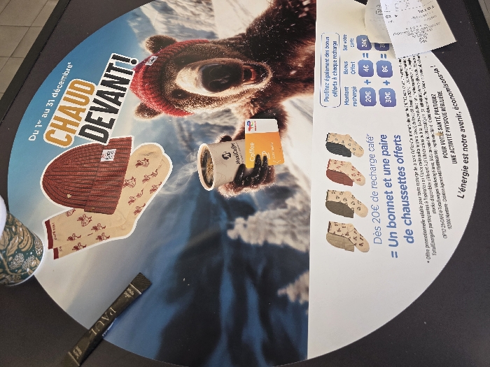 The image displays a circular promotional advertisement, approximately 30-40 cm in diameter, placed on a dark, flat surface, likely a countertop within a commercial establishment in France. The advertisement features a winter-themed graphic with a digitally rendered brown bear wearing a red knitted beanie, holding a white MaxiCoffee branded cup of steaming coffee. The bear is set against a blurred snowy mountain backdrop with conifer trees. Text in French reads "Du 1er au 31 décembre*", "CHAUD DEVANT!" (Warm Up Front!), and promotes an offer: "Dès 20€ de recharge café = Un bonnet et une paire de chaussettes offerts" (From €20 coffee top-up = A free beanie and a pair of socks). A red knitted beanie and a pair of light beige patterned socks are depicted above the bear. A bonus offer table details rewards for coffee recharges on a TotalEnergies branded card, showing "20€ + 4€ = 24€" and "30€ + 6€ = 36€". Below this, four pairs of patterned socks in various colors (light beige, green, red, dark gray) are illustrated. Fine print in French states the offer is valid at participating TotalEnergies stations in metropolitan France. The slogan "L'énergie est notre avenir, économisons-la!" (Energy is our future, let's save it!) appears at the bottom. To the upper left of the advertisement, a portion of a patterned paper cup is visible. To the left, a black rectangular sachet labeled "PAUL" rests on the dark surface. A crumpled white paper receipt partially overlaps the lower right section of the advertisement. The image itself does not provide the specific city of Urvillers, France, but indicates a TotalEnergies promotion occurring within metropolitan France.