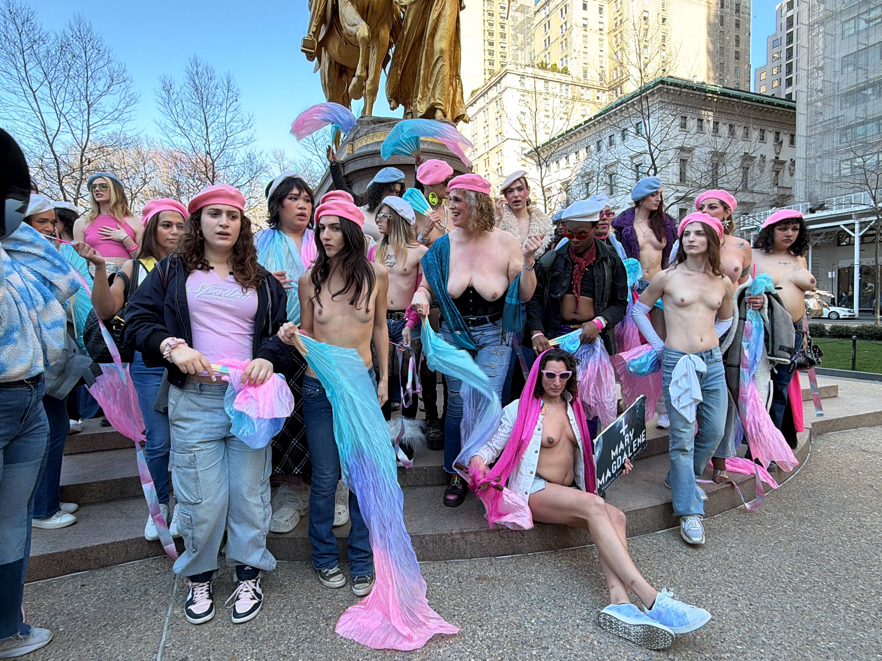 A group of people, mostly topless women, are gathered on a city street in front of a statue and a building in New York City, United States. Most of them are wearing jeans and pink hats. Some hold semi-transparent scarves in blue and pink colors. One person is sitting on the ground and holding a sign that reads "Mary Magdalene."