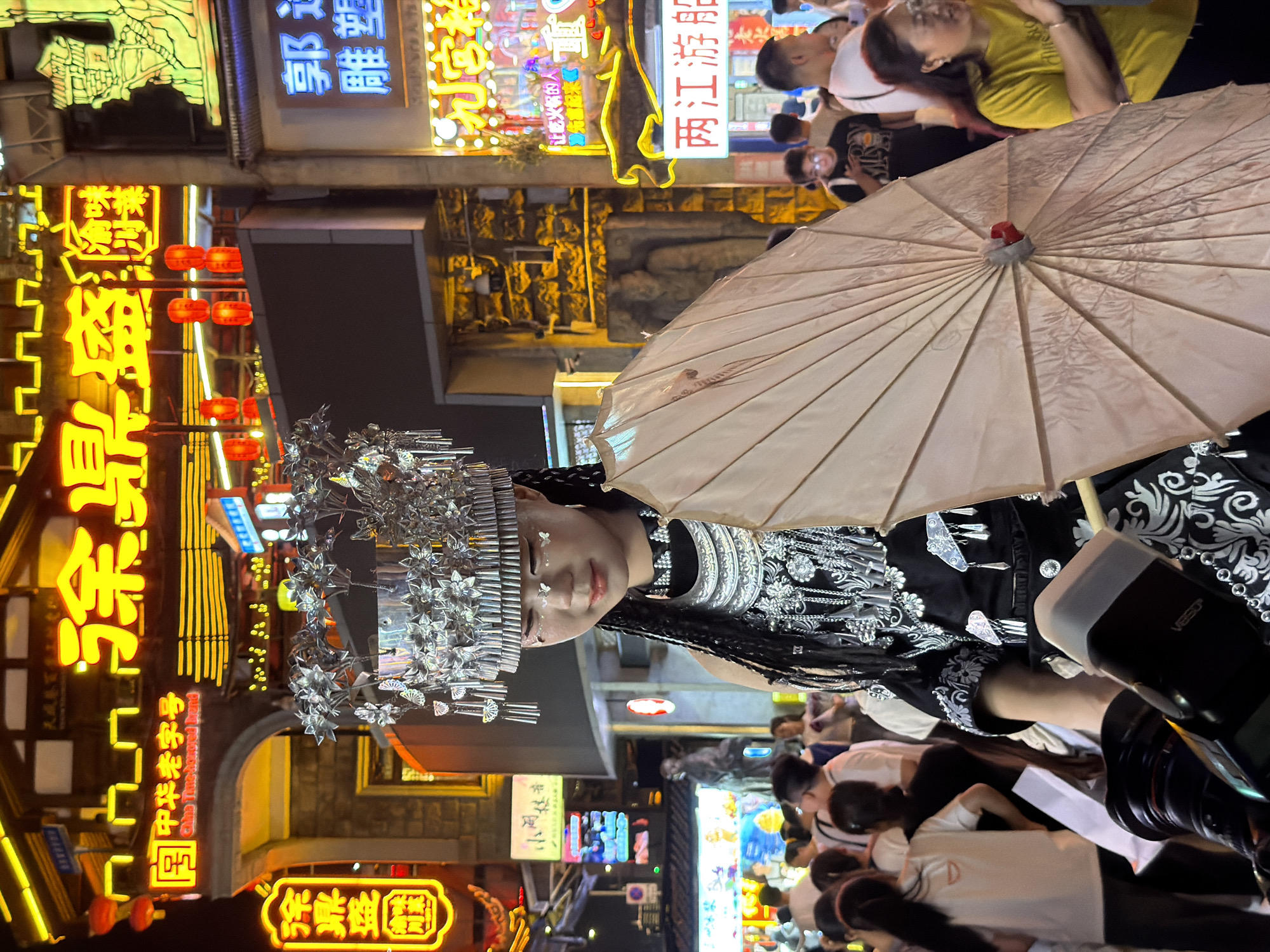 The image depicts a night scene in a vibrant, urban environment, likely a pedestrian street or market, with a focus on traditional Chinese culture. The prominent subject is a young woman dressed in elaborate traditional attire, including a large, ornate silver headdress adorned with intricate patterns and hanging ornaments. She has braids and is wearing a black garment with white embroidered designs. She is holding a light-colored, open umbrella, possibly made of paper, which partially obscures her lower body. Her face is visible, and she appears to be looking down and slightly to the side with a gentle expression. There are decorative elements on her face, resembling glitter or small decals.
In the background, the environment is brightly lit with numerous neon signs in Chinese characters, indicating shops and businesses. Some signs feature stylized depictions and traditional architectural elements. Red lanterns are also visible, adding to the festive atmosphere. The street appears to be crowded with people, suggesting a popular tourist or local gathering spot. Many individuals are visible in the background, some walking and others gathered in groups. The overall scene suggests a cultural festival, a traditional performance, or a lively market district at night.
The location is identified as Yuzhong District, China. This district is known for its historic areas and bustling commercial streets, which aligns with the visual cues in the image. The presence of traditional clothing, neon signage, and a crowd of people is characteristic of many vibrant urban centers in China, particularly those that blend modern commerce with cultural heritage. The lighting suggests it is nighttime, and the atmosphere is lively and engaging.