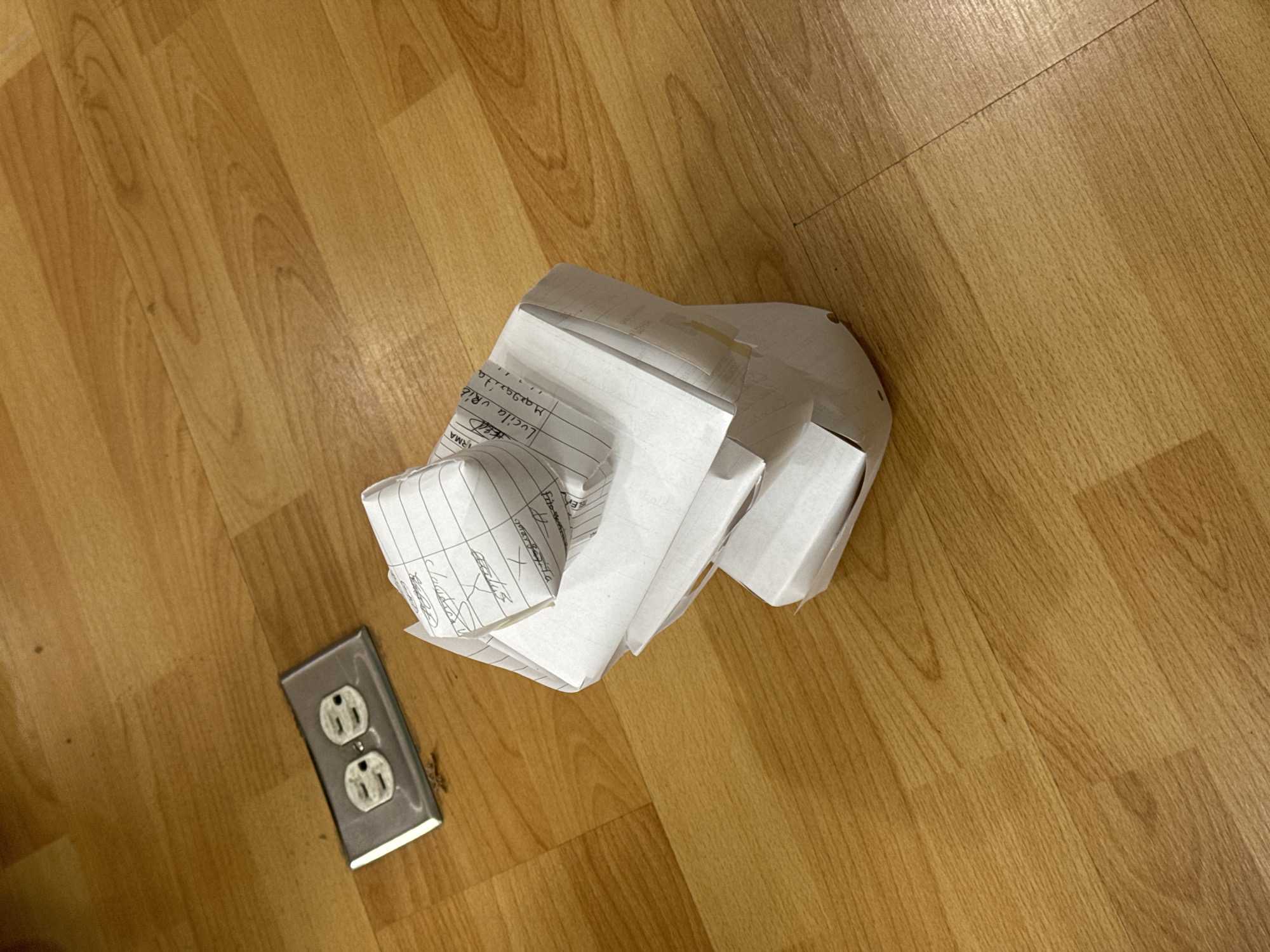 The image shows a section of a wooden floor. On the floor, there is a crumpled pile of white paper, some of which appears to have handwritten text. Beside the paper is an electrical outlet with two receptacles. The location is not identifiable as San Bernardino Tlaxcalancingo, Mexico, from the visual information provided.