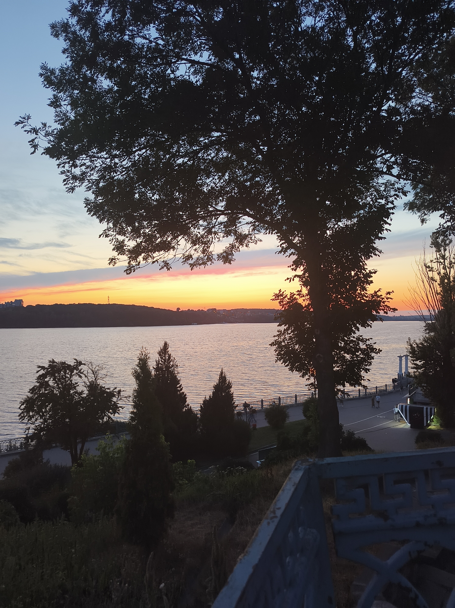 The image depicts a tranquil scene of a sunset over a wide body of water, likely Ternopil Pond, in Ternopil, Ukraine. The sky is painted with hues of orange, pink, and yellow, transitioning into a soft blue as it reaches the top of the frame. Silhouetted against the vibrant sky are the lush leaves and branches of a large tree in the foreground, casting a dark shadow over the scene.
Below the water, a well-maintained embankment lined with a decorative fence can be seen. A paved walkway runs along the embankment, where a few individuals are strolling, enjoying the evening ambiance. The embankment is dotted with trees and other vegetation, contributing to the natural beauty of the surroundings. In the distance, the silhouette of the city's buildings and structures can be discerned against the horizon, adding a touch of urban context to the otherwise natural panorama.
The overall impression is one of peacefulness and serenity, a perfect moment captured as day transitions into night. The interplay of light and shadow, the vibrant colors of the sunset, and the presence of nature and human activity create a harmonious and picturesque composition.