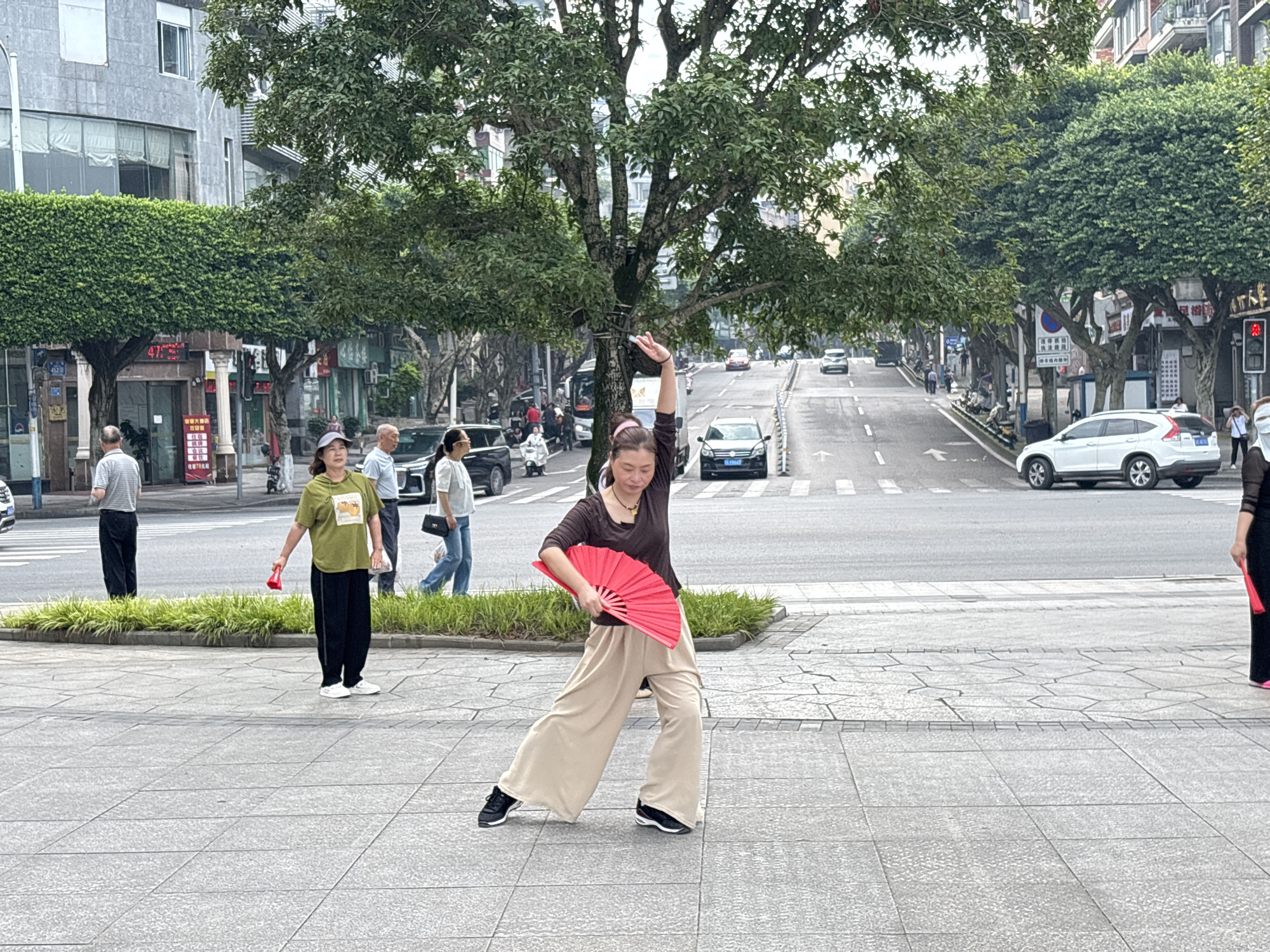 A woman in beige wide-leg pants and a brown t-shirt is captured mid-pose, holding a vibrant red fan. Her left arm is extended upward and bent at the elbow, while her right arm holds the fan open and angled across her body. She is performing some form of dance or exercise on a tiled pedestrian walkway. Behind her, a street curves uphill, with cars and buses visible. Trees with dense green foliage line the street, and buildings of varying heights form the urban backdrop. To her left, another woman in a green t-shirt and black pants is also holding a red fan, suggesting they might be practicing together. Several other individuals, including men and women, are walking or standing nearby, contributing to the bustling atmosphere of a city street in Jiangjin, China. The overall scene depicts a moment of cultural activity within an urban environment.