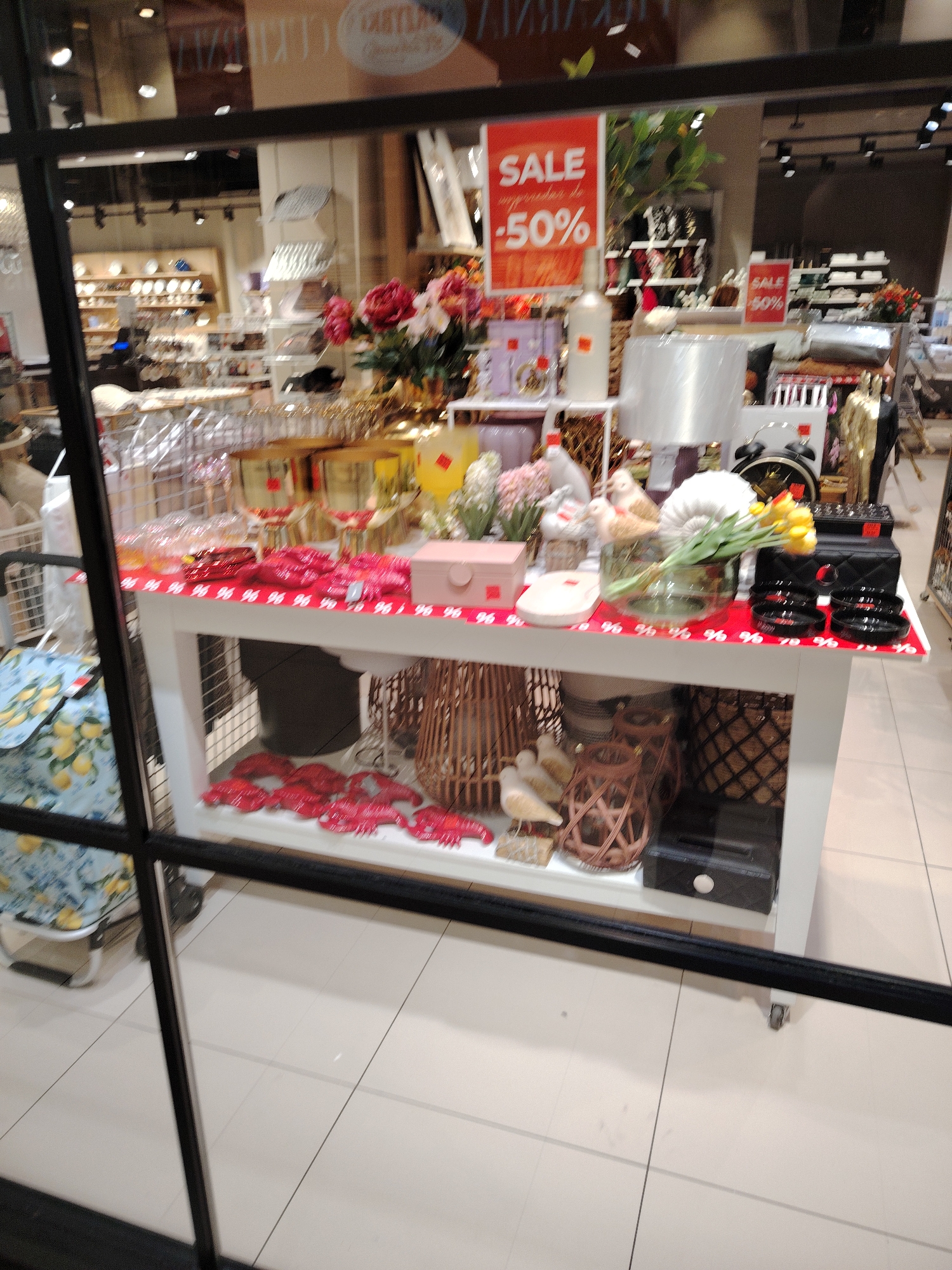 This is an eye-level, medium shot of a store window display. The display features a white table with various decorative items. On the tabletop, to the left, are several red lobster-shaped decorations. Next to these are pink boxes, and further to the right, a vase with yellow flowers, a stack of black bowls, and a quilted black bag. Behind the table, an assortment of merchandise is visible, including lamps, vases, and other decorative items.
A large red sign on the left side of the display reads "SALE -50%". A smaller red sign on the right also says "SALE 50%". The store appears to be a home decor or gift shop. The window is framed by thick black bars, suggesting it's a storefront. The background shows other shelves of products, indicating this is a retail environment. The floor tiles are visible in the foreground, and the overall lighting suggests it is daytime. The display is intended to attract customers by highlighting sale items and aesthetically pleasing products. No people are visible within the frame.