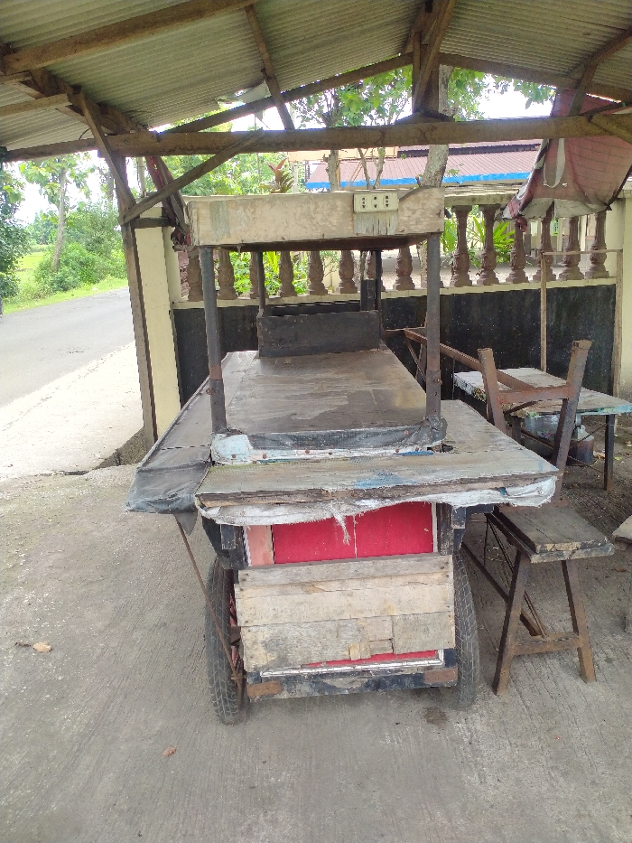 The image presents an empty, rustic street vendor's cart situated under a basic lean-to roof, constructed from corrugated metal sheets supported by wooden beams. The setting is outdoors, likely by a roadside, with a paved road and dense green foliage visible to the left.
The central subject is a well-used, mobile food cart, predominantly made of wood. It features a large, flat, dark-colored main counter surface. An elevated wooden structure above the main counter displays a row of three electrical outlets. A black, rectangular container or basin rests on the main counter. The lower part of the cart shows weathered wooden panels and sections painted in red. Two wheels are visible on the left side, indicating its function as a mobile stall.
To the right of the cart, a simple wooden stool with an A-frame base sits on the concrete ground. Further back, another small table or bench holds an inverted chair. In the background, a light-colored building with a decorative balustrade and a dark-painted lower section is partially visible behind trees.
The scene suggests daytime, with soft, diffused lighting indicating possibly overcast weather. No individuals are present, and there is no discernible activity taking place. The general appearance of the cart and its surroundings evokes a small, local business environment typical of places like Kecamatan Kebakkramat, Indonesia. There is no visible text.