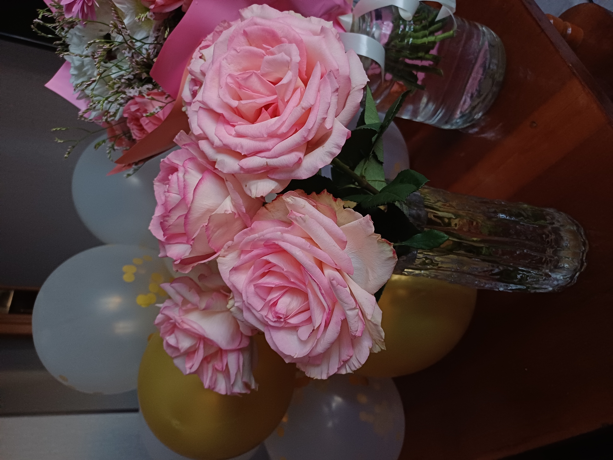A bouquet of light pink roses is displayed in a clear glass vase. The bouquet also contains smaller white flowers and green leaves. Several balloons, some light gray with yellow dots and others golden yellow, are arranged around the vase, suggesting a festive occasion. The setting appears to be indoors, possibly a table, with a wooden surface visible in the background. The lighting is soft, indicating it might be daytime or a well-lit interior. The overall mood is celebratory and cheerful. There is no visible text.