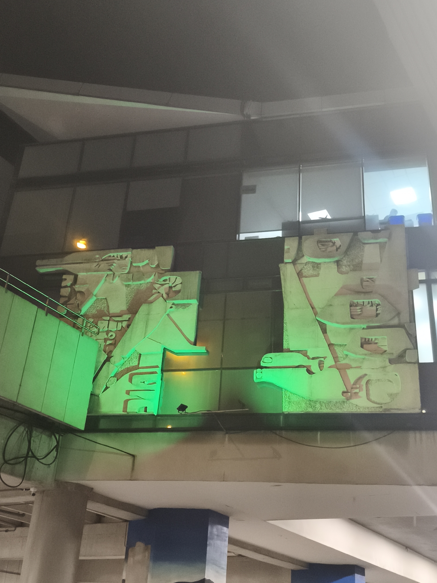 Here is a detailed description of the image:
The image shows a building interior in Guatemala City, Guatemala. The architecture is modern, with gray, white, and blue hues in the lower sections. Large panels with stylized, low-relief artwork are illuminated with bright green light. The artwork appears to be of Mayan or other indigenous Guatemalan origin, possibly replicas of historical carvings. Behind the panels are windows into what appears to be an office space, with lights on and blurry shapes of people visible inside.