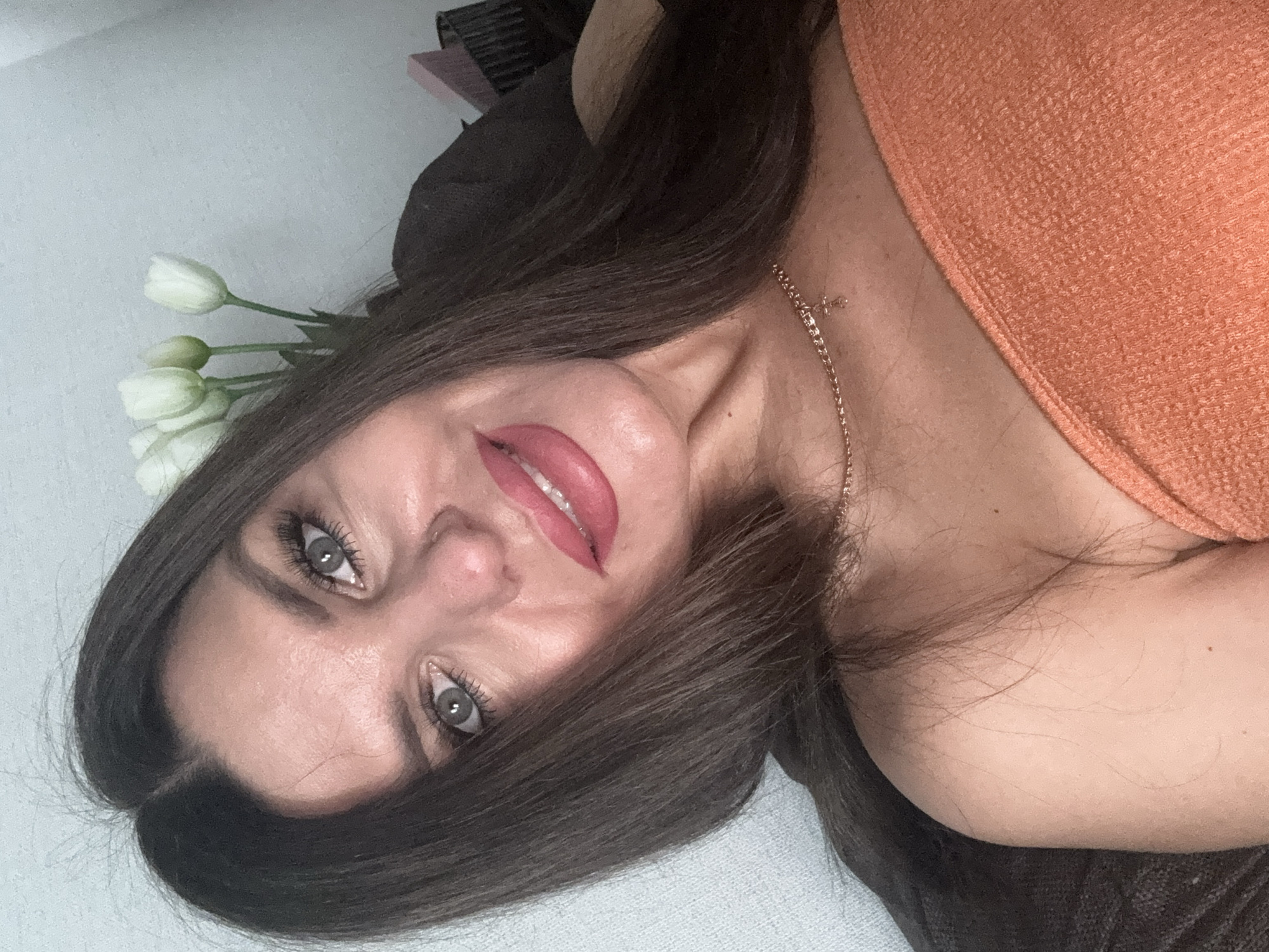 A medium shot shows a woman from the chest up, looking directly at the camera. She has long, dark brown hair that falls around her shoulders and is parted in the middle. Her eyes are a light greyish-blue, with dark eyelashes and eyebrows. She is wearing a coral-colored strapless top and a delicate gold chain with a small cross pendant around her neck. Her lips are full and painted a bright pink lipstick. The background is softly blurred, with a wall and what appear to be white tulips in the upper right corner. The lighting is warm, creating a slight glow on her skin. The scene appears to be indoors, possibly in a home setting. There is no indication of the location being Chaikovsky, Russia.