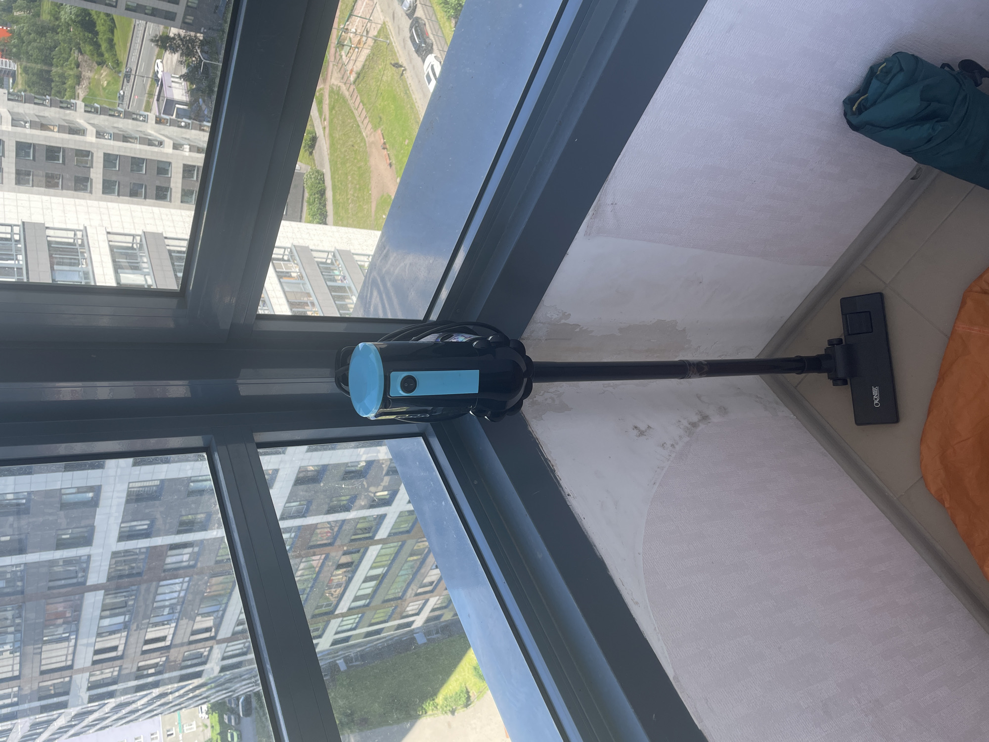 In the city of Sinyavino, Russia, a vertical shot shows a black and blue upright vacuum cleaner positioned in the corner of a room with large windows overlooking a dense urban landscape. The vacuum, with a blue top and black body, stands upright on a beige tiled floor. Its brush attachment is visible at the base. The room's wall is covered with a textured white wallpaper, showing some discoloration and peeling at the corner. To the right, a rolled-up dark green sleeping bag leans against the wall. The window reveals multiple high-rise buildings, some with a grey and white facade, and others with darker detailing. Green trees are interspersed between the buildings, and a road with parked cars is visible in the distance. The sky appears overcast.