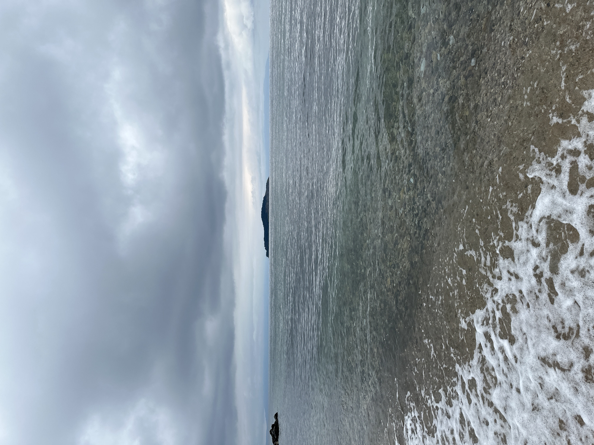 The image depicts a shoreline scene in Ολυμπιάδα, Greece. A body of water occupies the right side of the frame, with gentle waves washing onto a pebble beach. The water appears clear, revealing pebbles beneath the surface. On the left, dark, overcast skies fill the frame, creating a muted, gray atmosphere. In the distance, a small landmass is visible, possibly an island or peninsula. No people are visible in the image.