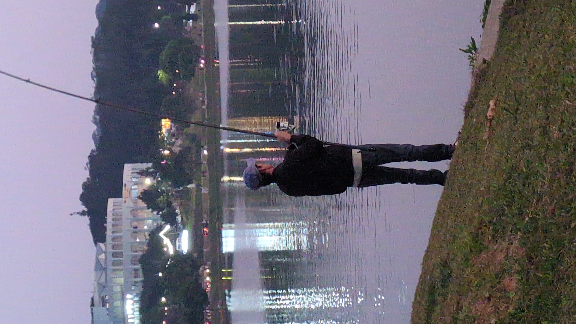 A man in a dark jacket and blue baseball cap is fishing in a lake in Da Lat, Vietnam. He stands on a grassy bank, holding a fishing rod, with a large building and street lights reflected in the water. The scene appears to be at dusk or night. The background includes trees and a hillside.
