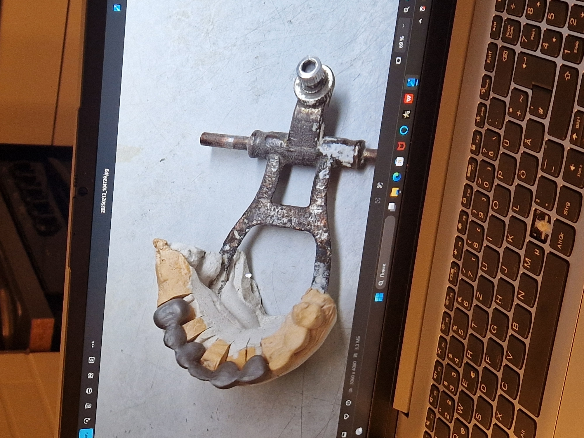 The image shows a laptop screen displaying a photograph of a dental mold with a metallic apparatus attached. The dental mold appears to be made of plaster and wax, with a row of teeth-like structures. The metallic apparatus is a complex, rusty structure with levers and a screw mechanism. The laptop is open, and the keyboard is visible in the foreground. The background appears to be an indoor setting with a neutral-colored surface. There are no people visible in the image. The filename on the screen suggests the image was taken on February 13, 2025. There is no information in the image to indicate the location in the city of Leimen, Germany.