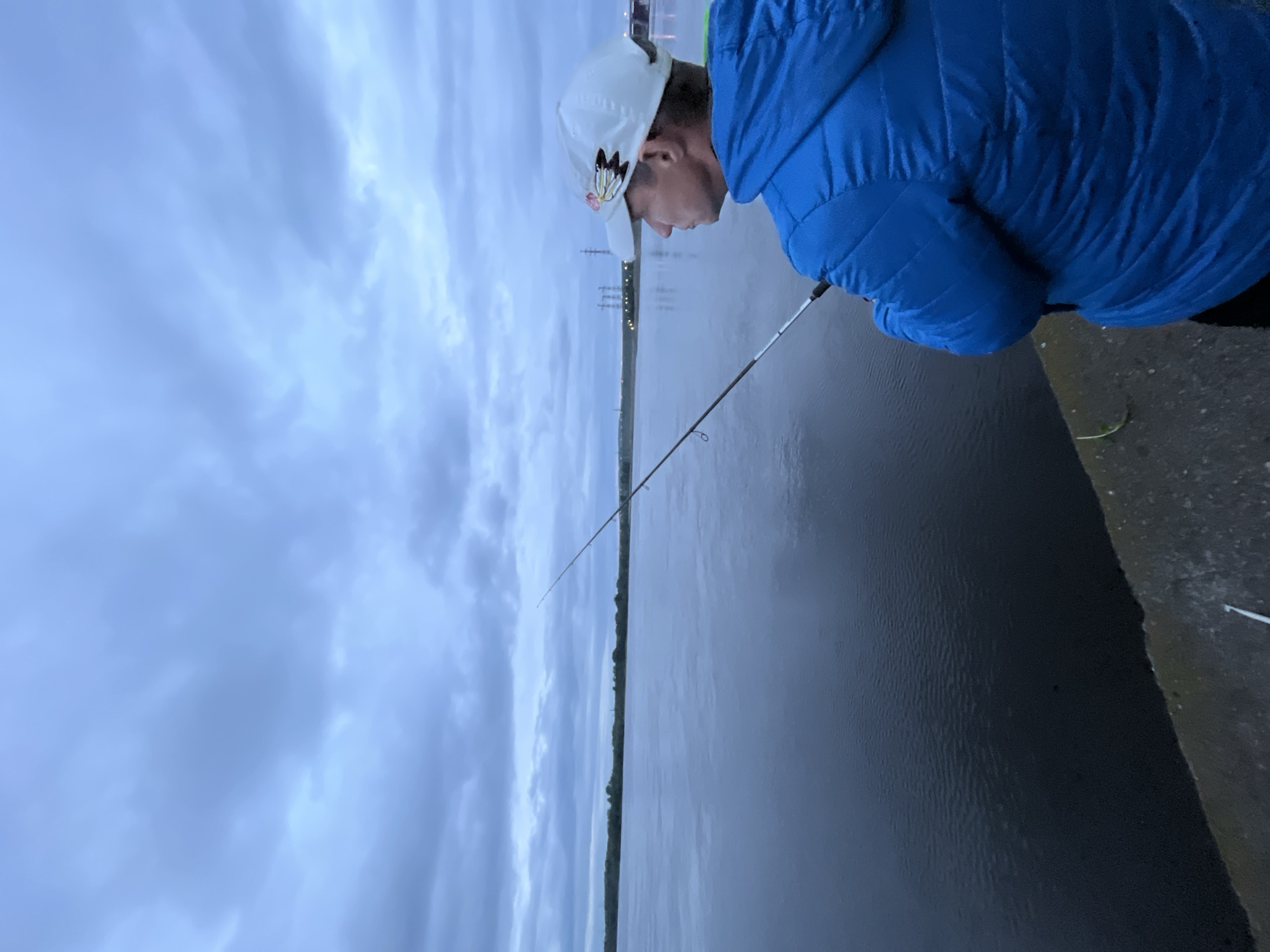 An outdoor scene in Noviy, Russia, depicts a single individual fishing by a large body of water under an overcast sky. The person, a male, is viewed from the back and right side, wearing a blue quilted hooded jacket and a white baseball cap adorned with a multi-colored embroidered feather design on the right temple. He holds a fishing rod angled downwards towards the water. The individual stands on a grey concrete embankment or wall in the immediate foreground. The expansive body of water, likely a river or lake, appears dark blue-grey and exhibits subtle surface ripples. A green tree-lined horizon is visible across the water. In the distant right, several industrial-style power line pylons or similar structures are silhouetted against the horizon, with some faint light reflections. The sky above is entirely covered with grey-blue clouds, indicating an overcast condition, likely at dawn or dusk due to the diffused low light.