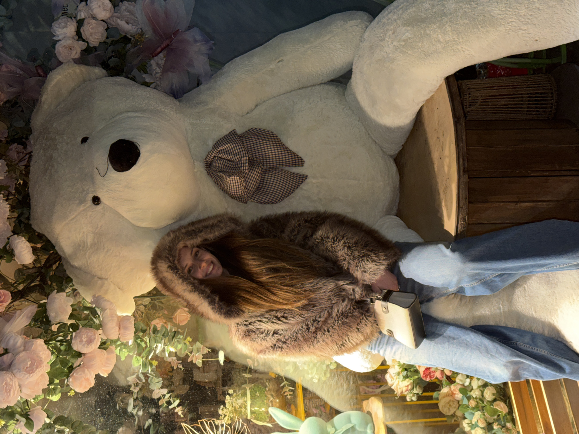 A woman in a fur coat and jeans is lying on top of a large, white teddy bear decorated with a plaid bow. The teddy bear is seated in a wooden crate and surrounded by artificial flowers. The scene is likely a themed photo opportunity in Sofia, Bulgaria. The woman is smiling and appears to be posing for a photo.