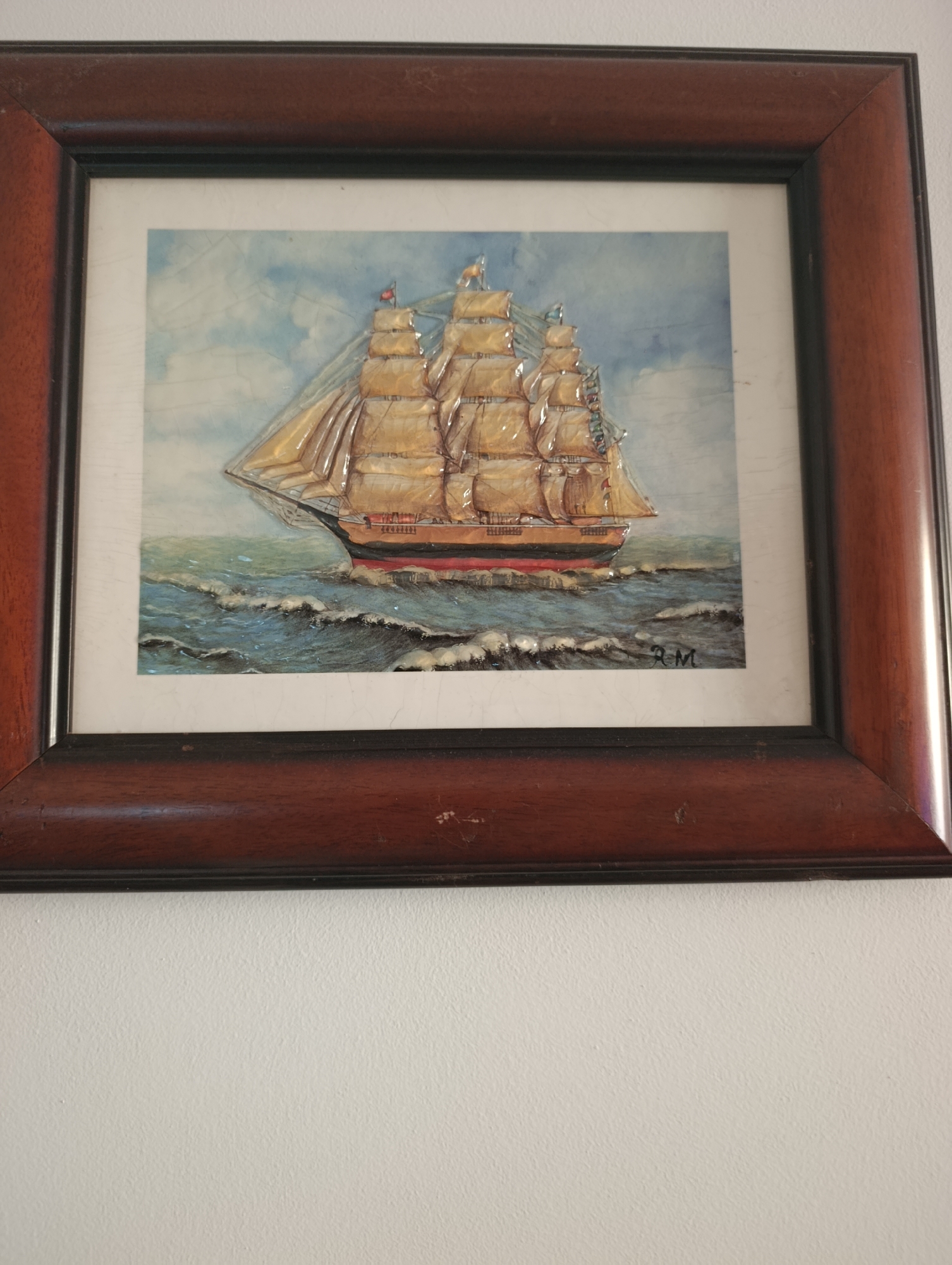 A framed painting of a tall ship sailing on a wavy sea is hung on a white textured wall. The painting depicts a three-masted ship with full sails, rendered in warm tones of brown, gold, and red. The sky is a light blue with wispy white clouds. The signature "RM" is visible in the lower right corner of the painting. The frame is made of dark wood with a subtle grain pattern. The painting is hung at eye level, with the top of the frame approximately at the midpoint of the wall.