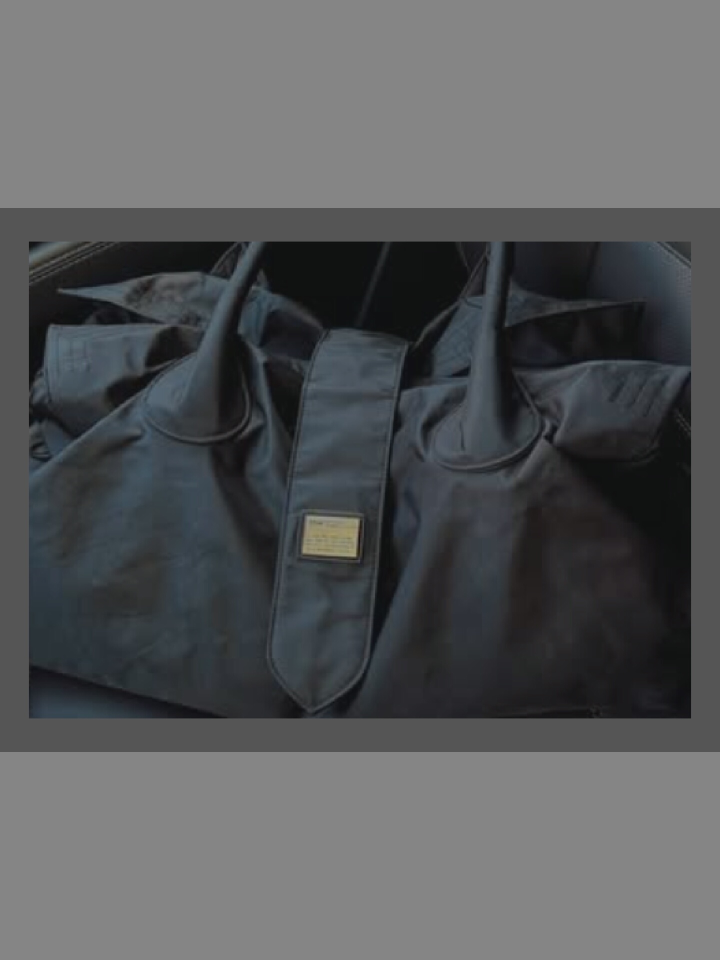 A dark, textured duffel bag rests on a dark, upholstered surface, possibly a car seat. The bag is made of a matte, woven material. A prominent feature is a central strap with a small, rectangular metal tag. The tag appears to have some text engraved on it, but it is too small to be legible. The bag has two handles that loop over the top. The lighting is dim, casting subtle shadows and highlighting the texture of the bag's fabric. The background is blurred, suggesting the focus is entirely on the bag.
