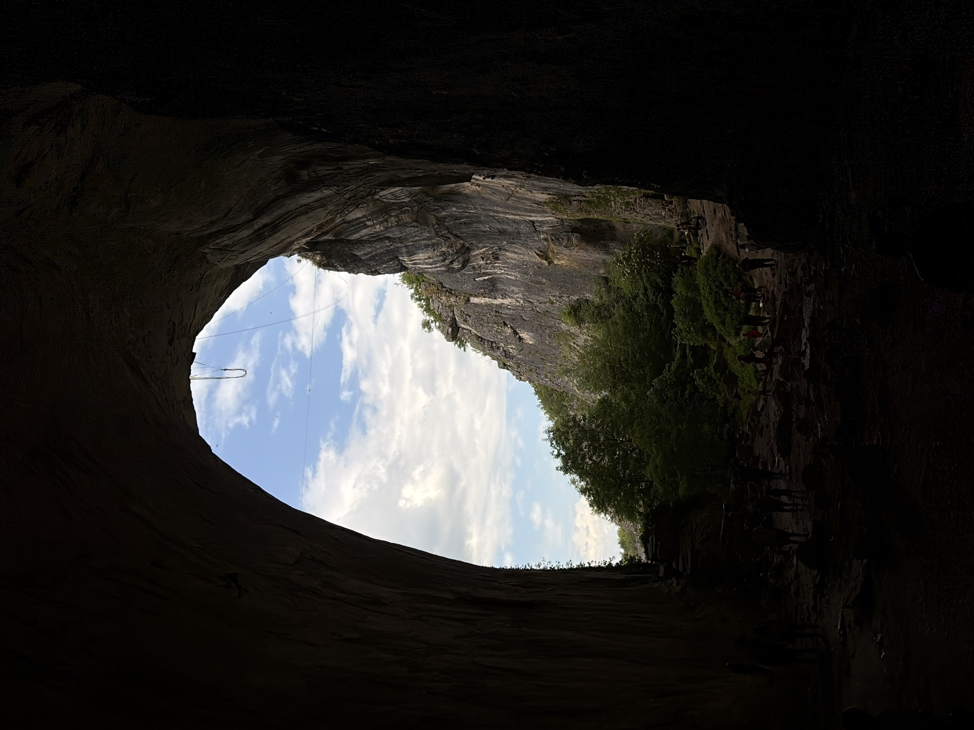 View from inside a dark cave opening looking outward toward a bright sky. The cave opening is large and oval-shaped. Outside, layered rock formations with green vegetation are visible. A group of indistinct figures appears to be standing or walking along a path at the top of the cave entrance. A thin wire spans across the top opening of the cave. The location is Unknown, Unknown.