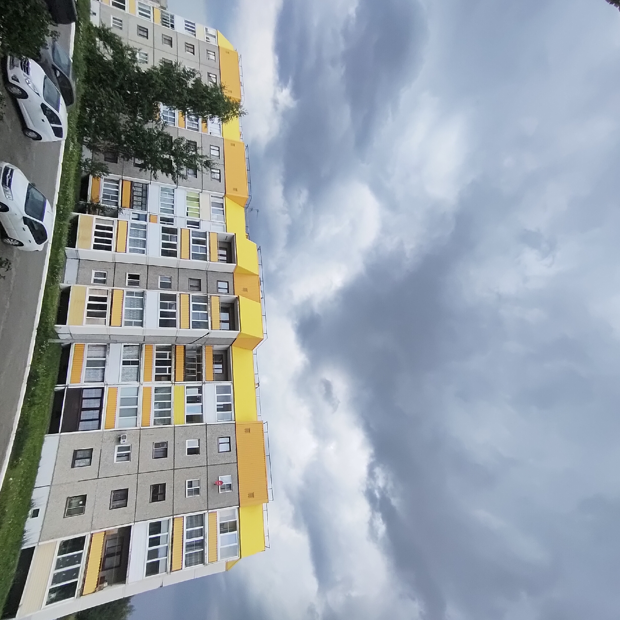 This is a wide-angle, eye-level outdoor photograph of a multi-story apartment building with a yellow roofline and grey concrete facade, located in the city of Марковский, Russia. The building is curved and appears to be a typical Soviet-era residential block, with numerous windows and balconies. There are trees on the left side of the building, and a parking lot with several cars in front of it. The sky is overcast with dark, heavy clouds, suggesting impending rain or a storm. The overall impression is a mundane urban residential scene under a dramatic sky.
