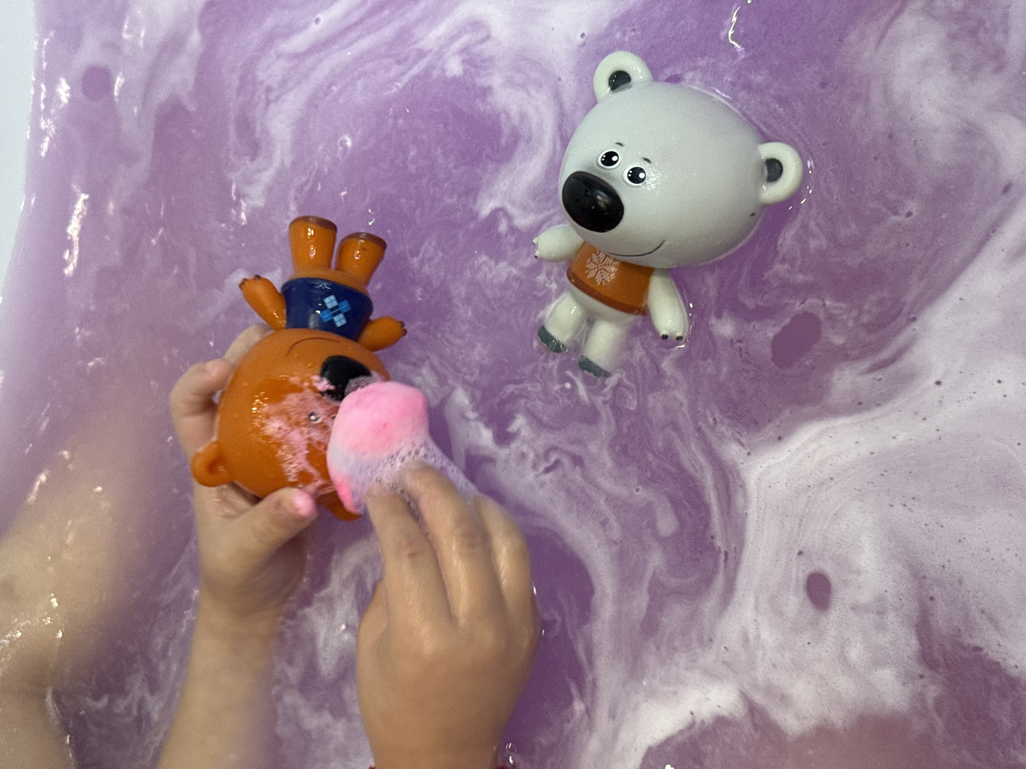 The image shows a close-up of a person's hands holding an orange toy bear in purple bathwater. The bear is wearing a blue outfit with white squares. A pink sponge is near the bear. In the lower right portion of the image, a white toy bear with a black nose and eyes, wearing an orange outfit with a snowflake pattern, floats in the water. The bathwater is a swirling mix of purple and white foam. No specific location in Lyubertsy, Russia, can be determined from this image.
