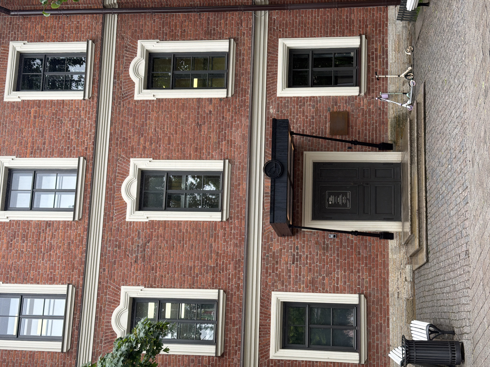 The image depicts the exterior of a red brick building with white decorative trim around its windows. The building has multiple stories, with several windows visible on each level. The windows are dark-framed and appear to be multi-paned. On the ground floor, there is a dark-colored entrance with a small canopy above it. To the left of the entrance, there are two white benches and a black trash can. The ground is paved with cobblestones. To the right of the entrance, two scooters are parked on the cobblestone pavement. The building is located in Saint Petersburg, Russia.