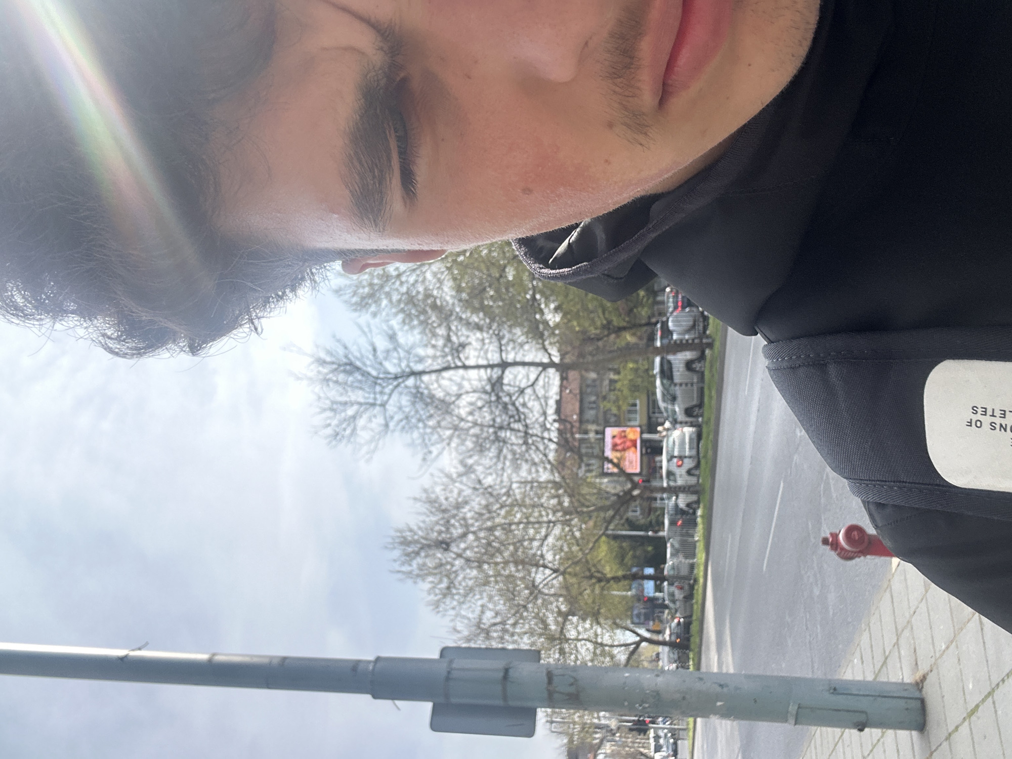 The image is taken in Plovdiv, Bulgaria, from a low-angle, tilted perspective. A portion of a person's face, including the forehead, eye, nose, and mouth, occupies the upper part of the frame. The person has dark hair and light skin. They are wearing a black hoodie or jacket.
In the background, a street scene unfolds. Cars are parked along the side of the road. There are trees with bare branches, suggesting either late autumn or early spring. There is also a billboard with a red image on it.
In the foreground, a grey metal pole with attachments is visible, possibly a street sign or lamp post. A red fire hydrant is also visible on the sidewalk. The sidewalk is paved with light-colored square tiles. The sky appears bright and slightly overcast.