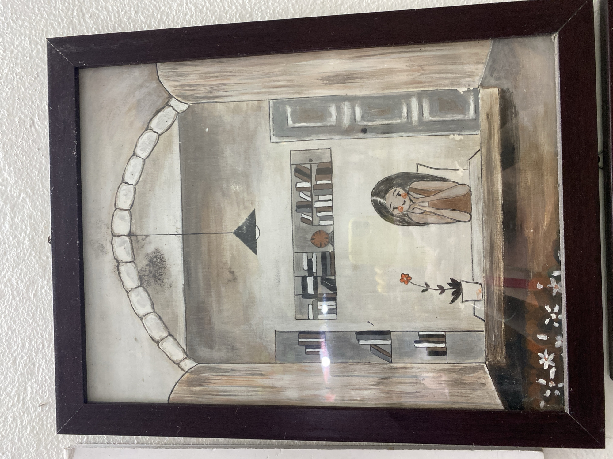 A framed painting depicts an interior scene with a young girl sitting at a desk. The girl has long, dark hair and is wearing a brown sleeveless dress. Her hands are clasped under her chin, and she has rosy cheeks and a gentle smile. In front of her on the desk is a potted plant with a single orange flower. Behind the girl, bookshelves are visible, filled with books. To the right, a closed door is painted. Above the desk, a hanging lamp is suspended from the ceiling. The scene is framed by architectural elements resembling an archway. The bottom of the painting shows a depiction of small white flowers. The painting is hanging on a textured white wall. The exact location in Đà Nẵng, Vietnam, is not specified within the image itself.