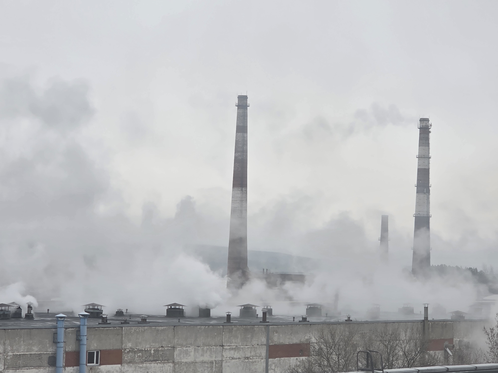 The image depicts a scene in Chaikovsky, Russia, showing industrial facilities releasing significant amounts of smoke and steam into the atmosphere. Visible are several smokestacks, some with distinct red and white stripes, rising above the rooftops of large industrial buildings. The air is thick with smoke and steam, creating a hazy, overcast environment. The architectural style is typical of industrial areas, with simple, utilitarian structures. The scene suggests heavy industrial activity and potential environmental pollution.