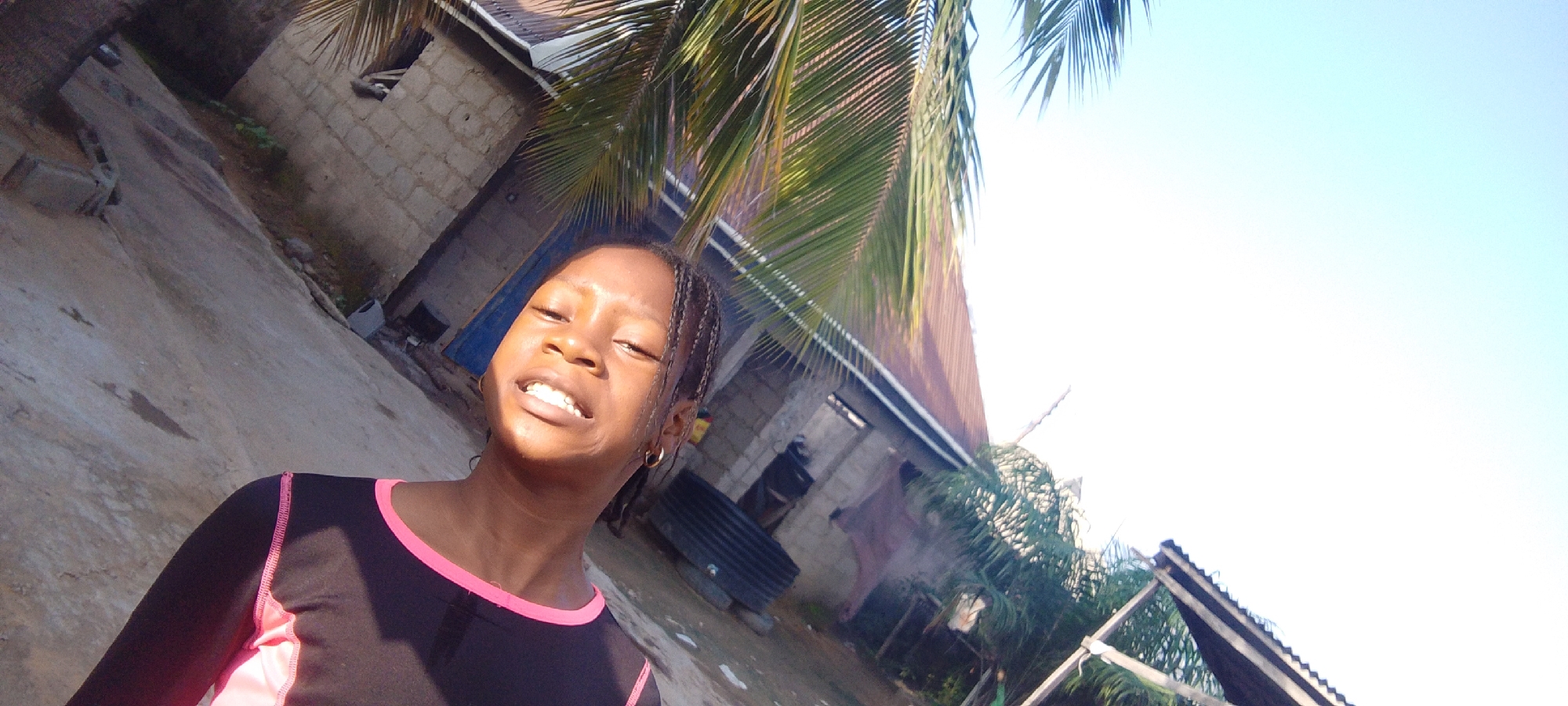 A young Black girl with braided hair and wearing a black long-sleeved shirt with pink trim smiles at the camera. She has gold hoop earrings on. She is outdoors in front of a building. The building appears to be made of cinder blocks and has a corrugated metal roof. There are palm fronds in the foreground and background, suggesting a tropical or subtropical location. The ground is dirt and concrete. The sky is clear and blue. The image is a selfie, as indicated by the angle and the fact that the girl is the primary subject.