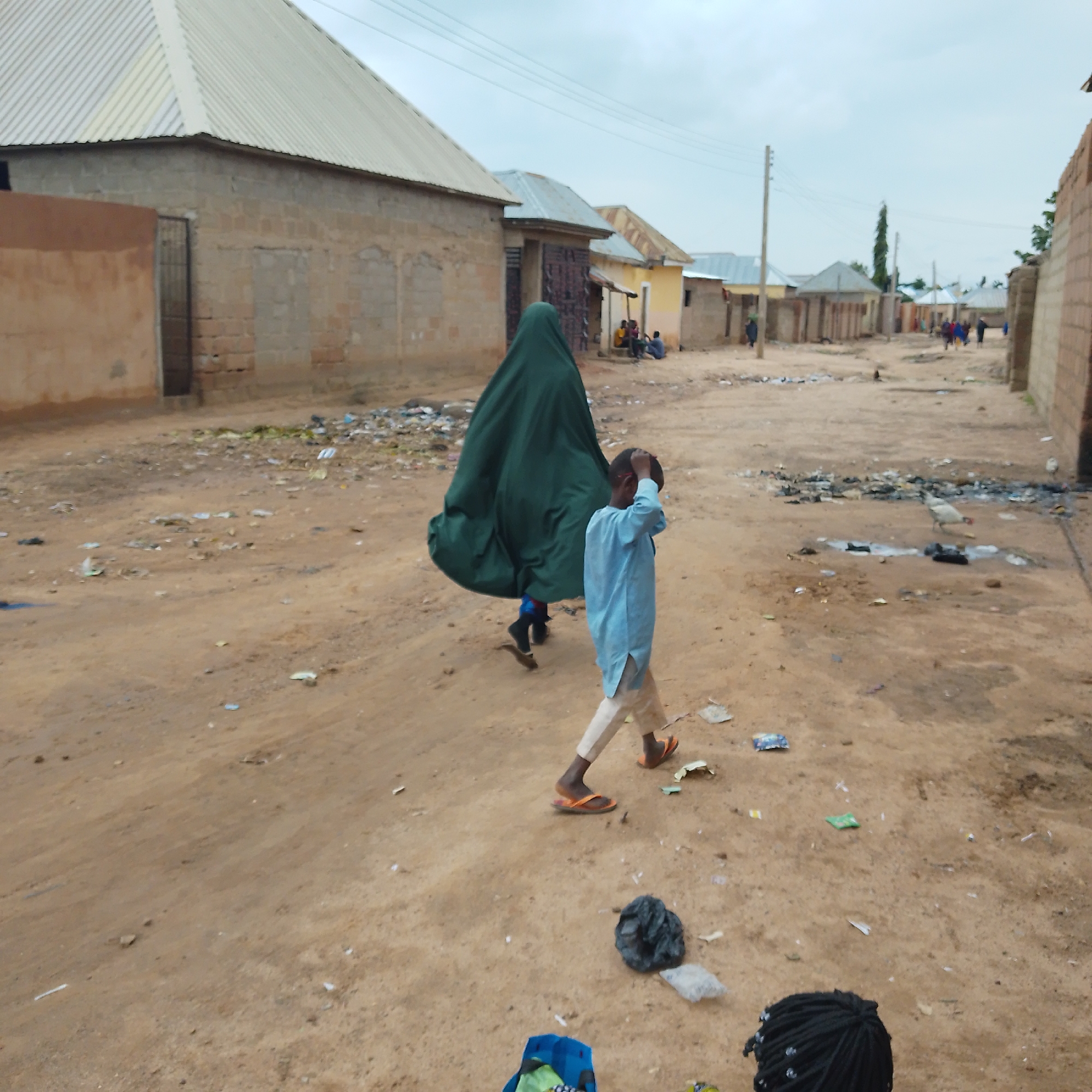 The image depicts a dusty street in Pantami, Nigeria, with scattered debris and some structures in the background. In the foreground, a young boy wearing a light blue tunic and white trousers walks from right to left, with his right hand touching his head. Behind him, a person covered in a large, dark green fabric walks in the same direction. The ground is dirt with various pieces of litter, including plastic bags and wrappers. In the middle ground, several people are seated outside a building. Further down the street, more structures and some distant figures are visible. The sky is overcast.