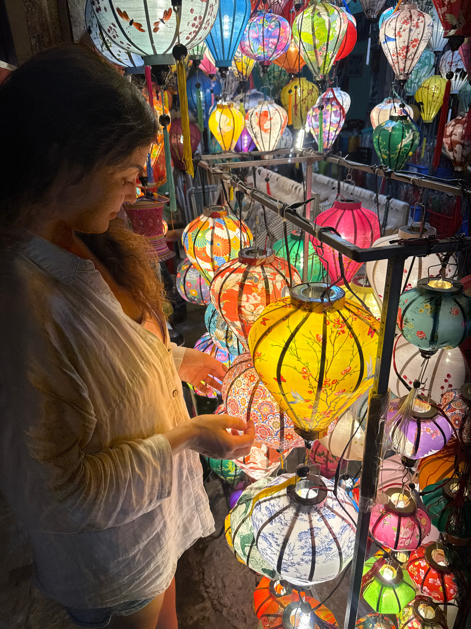 A woman with long, wavy brown hair is inspecting a stall filled with colorful, illuminated paper lanterns in Hoi An, Vietnam. She is wearing a light-colored, loose-fitting button-down shirt and denim shorts. The lanterns are of various sizes and designs, featuring floral patterns, birds, and butterflies. The stall is densely packed with lanterns, creating a vibrant and festive atmosphere. The lighting is dim, with the lanterns providing the primary source of illumination, casting a warm glow on the scene. The background is out of focus, but appears to be a market or street scene.