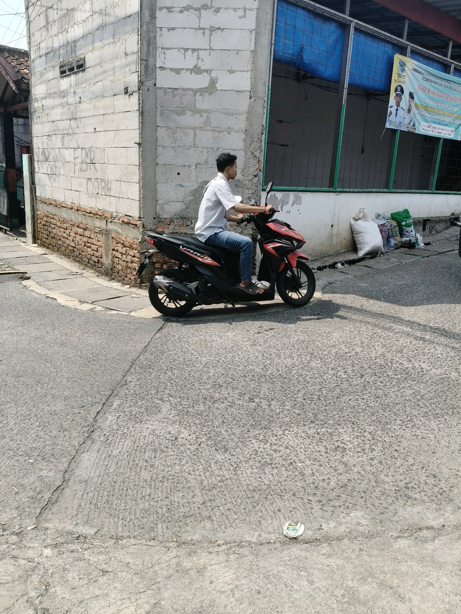 A man wearing a white short-sleeved shirt and blue jeans rides a red and black Honda Vario scooter on a cracked asphalt road in South Tangerang, Indonesia. The scooter is positioned in the center of the frame, facing left. Behind the scooter, a building with a white brick wall and a blue-gridded window is visible. A banner with Indonesian text and images of men in uniform hangs on the building. To the right of the scooter, several large sacks are stacked near the building's foundation. The foreground shows the textured asphalt road with tire tracks and shadows.