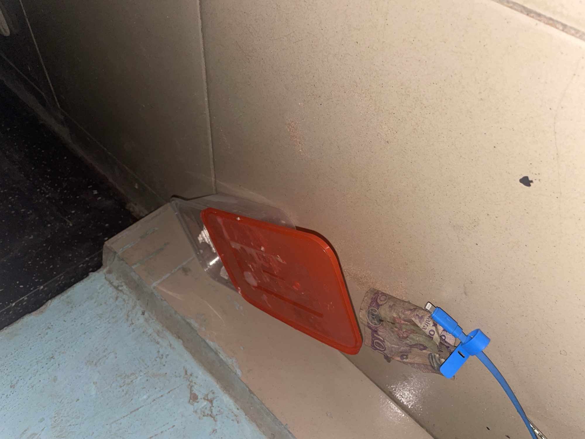 In a corner of a room in Jalingo, Nigeria, a red plastic lid rests on the floor next to a crumpled banknote and a blue charging cable. The banknote, appearing to be a 100 unit denomination, is partially obscured by the cable. The charging cable, with a white connector, is secured with a blue plastic tie. The floor is tiled, and a light blue wall forms a backdrop to the scene, with darker shadows in the background suggesting a poorly lit area. The overall environment appears somewhat dusty.