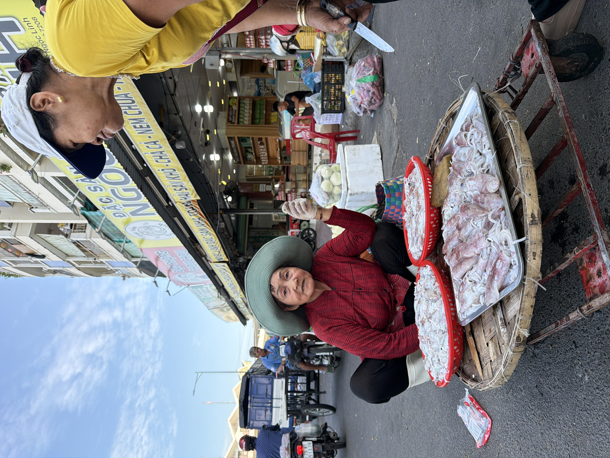 The image depicts a street scene in Nha Trang, Vietnam, likely near a market. In the foreground, a woman wearing a large, wide-brimmed hat and a red checkered shirt is crouching beside a cart. On the cart, there are two red baskets and a metal tray filled with raw squid. A woman in a yellow shirt and a white baseball cap stands to the right, holding a knife. In the background, several people are on motorcycles and a motorized tricycle, with shops and buildings lining the street. The sky is cloudy.