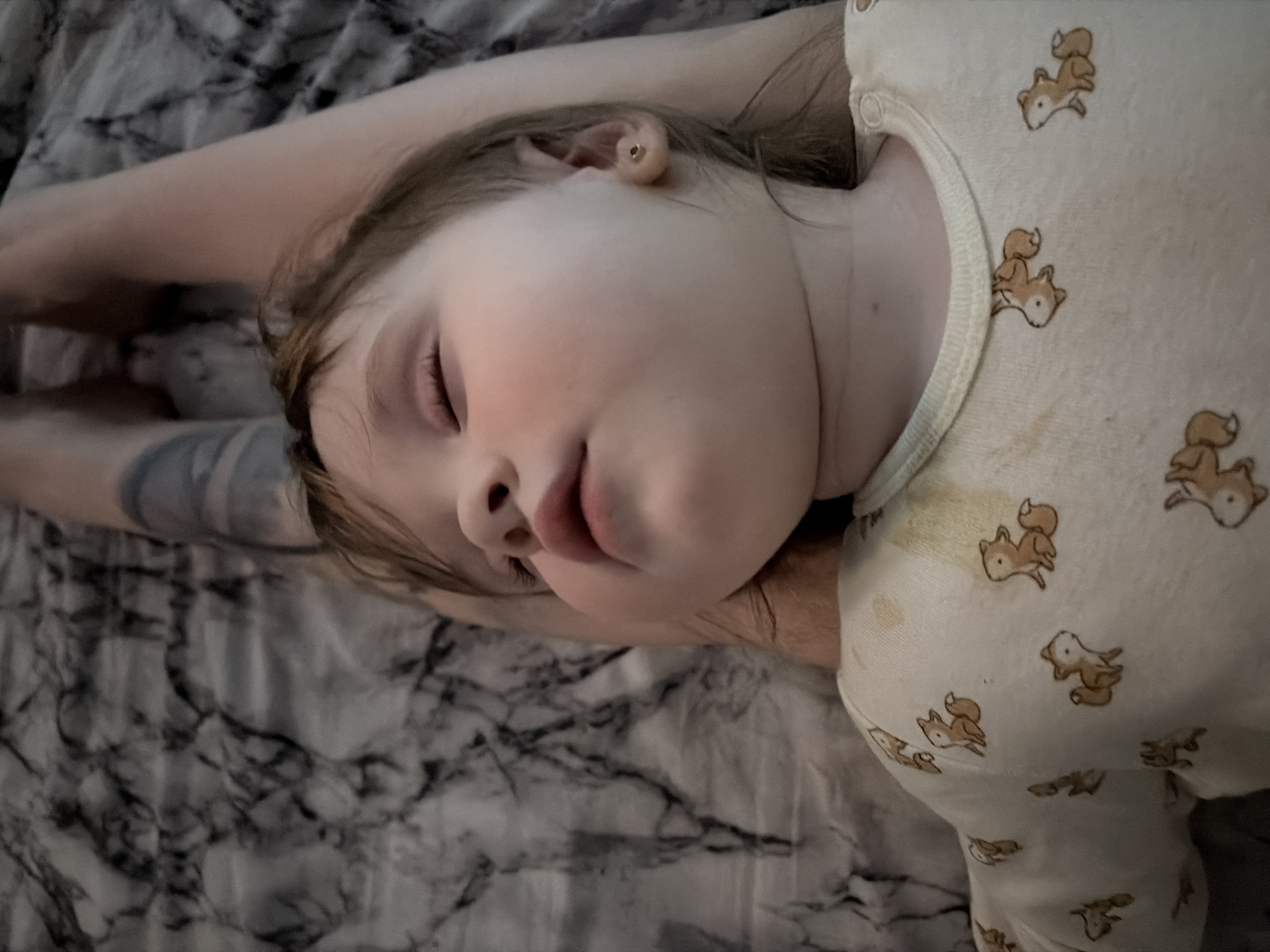 A child sleeps on a bed. The child is lying on their side, facing the camera. They are wearing a light-colored onesie patterned with foxes. An adult's arm, tattooed, is visible behind the child's head. The bedding has a marbled grey pattern. The location is undetermined.