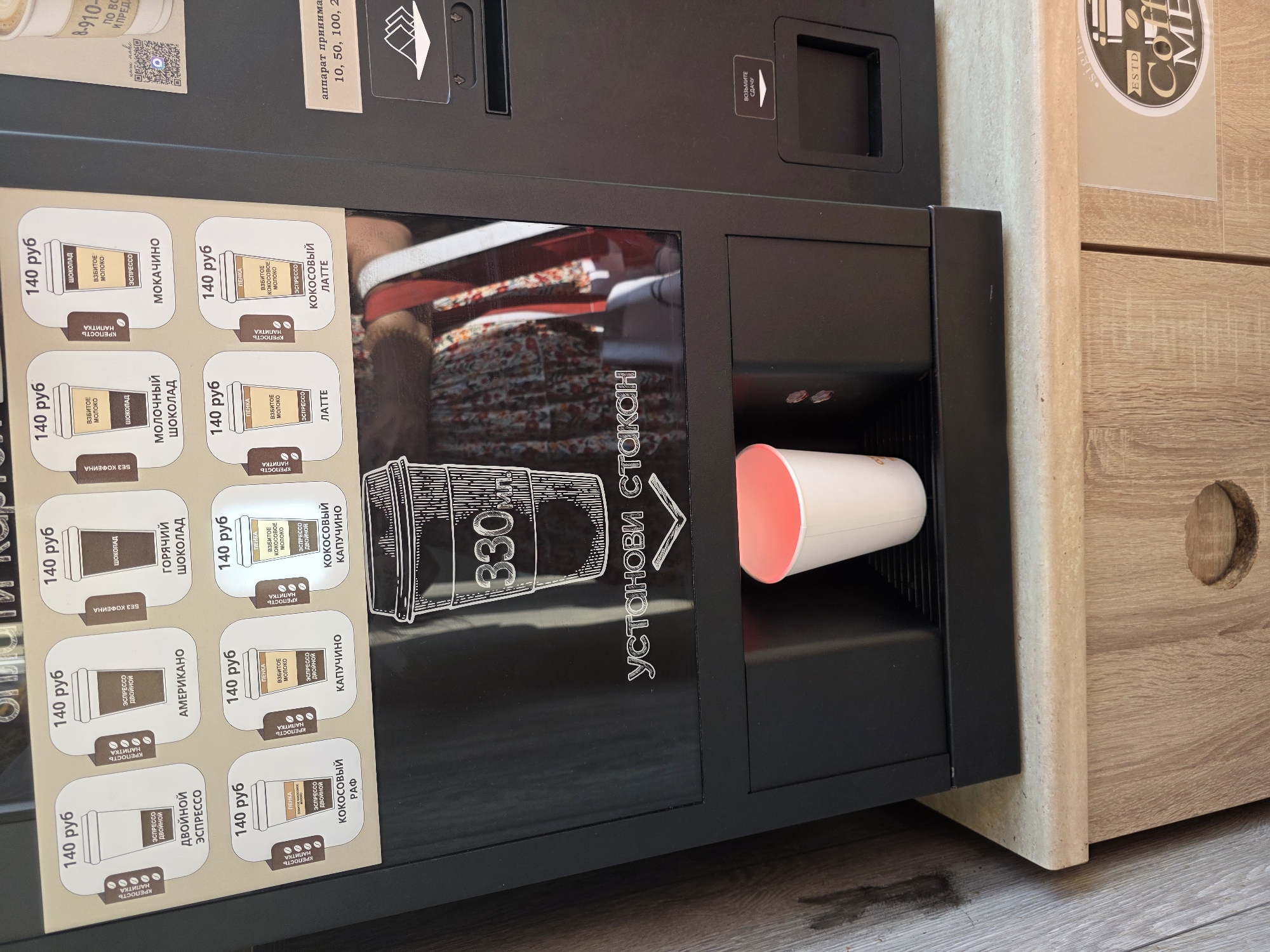 This is a close-up, eye-level, indoor shot of a black coffee vending machine. The machine's upper section displays various coffee options with their prices in Russian Rubles. Each option features an illustration of a cup and text indicating the drink's name, such as "ДВОЙНОЙ ЭСПРЕССО" (Double Espresso), "АМЕРИКАНО" (Americano), "ГОРЯЧИЙ ШОКОЛАД" (Hot Chocolate), "МОКАЧИНО" (Mocha), "КАПУЧИНО" (Cappuccino), and "КОКОСОВЫЙ ЛАТТЕ" (Coconut Latte). Below the menu, a large graphic of a 330ml cup is visible, with the text "УСТАНОВИ СТАКАН" (Place Cup) below it. A white paper cup with a pink rim is positioned in the dispensing area of the machine, partially filled with a pink beverage. To the right of the cup dispenser, a slot for inserting bills is visible, and below it, a coin return slot is labeled "ВОЗЬМИТЕ СДАЧУ" (Take Change). The machine is situated on a countertop made of a light-colored, textured material, with a wooden cabinet beneath it. A sticker on the lower right side of the cabinet features a circular logo with text that is partially obscured but includes "Coffee ME". The background consists of a textured, light-colored wall. There are no people clearly visible in the frame, though a reflection in the glass surface of the machine suggests a person wearing a patterned dress is standing behind it. The environment appears to be a public space, possibly a cafe or a rest area. There is no information in the image to place it in the city of Sochi, Russia.
