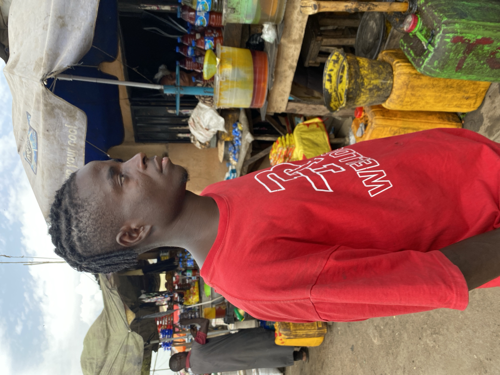 The image shows a young Black man wearing a red t-shirt with white lettering. He is standing in what appears to be an outdoor market stall in Bukuru, Nigeria. Behind him, the stall is stocked with various items, including bottled drinks, yellow and green plastic containers, and some white sacks. The scene suggests a typical market environment with goods for sale. The man's expression is neutral, and his gaze is directed slightly upwards and to his left.
The lighting and bright colors of the merchandise indicate it is daytime. The presence of the market stall and the variety of goods point to a commercial activity. The background shows more stalls and possibly a sky with clouds, suggesting an outdoor setting. The red t-shirt the man is wearing has white lettering that reads "WELLO". The overall impression is a snapshot of daily life in a Nigerian market.