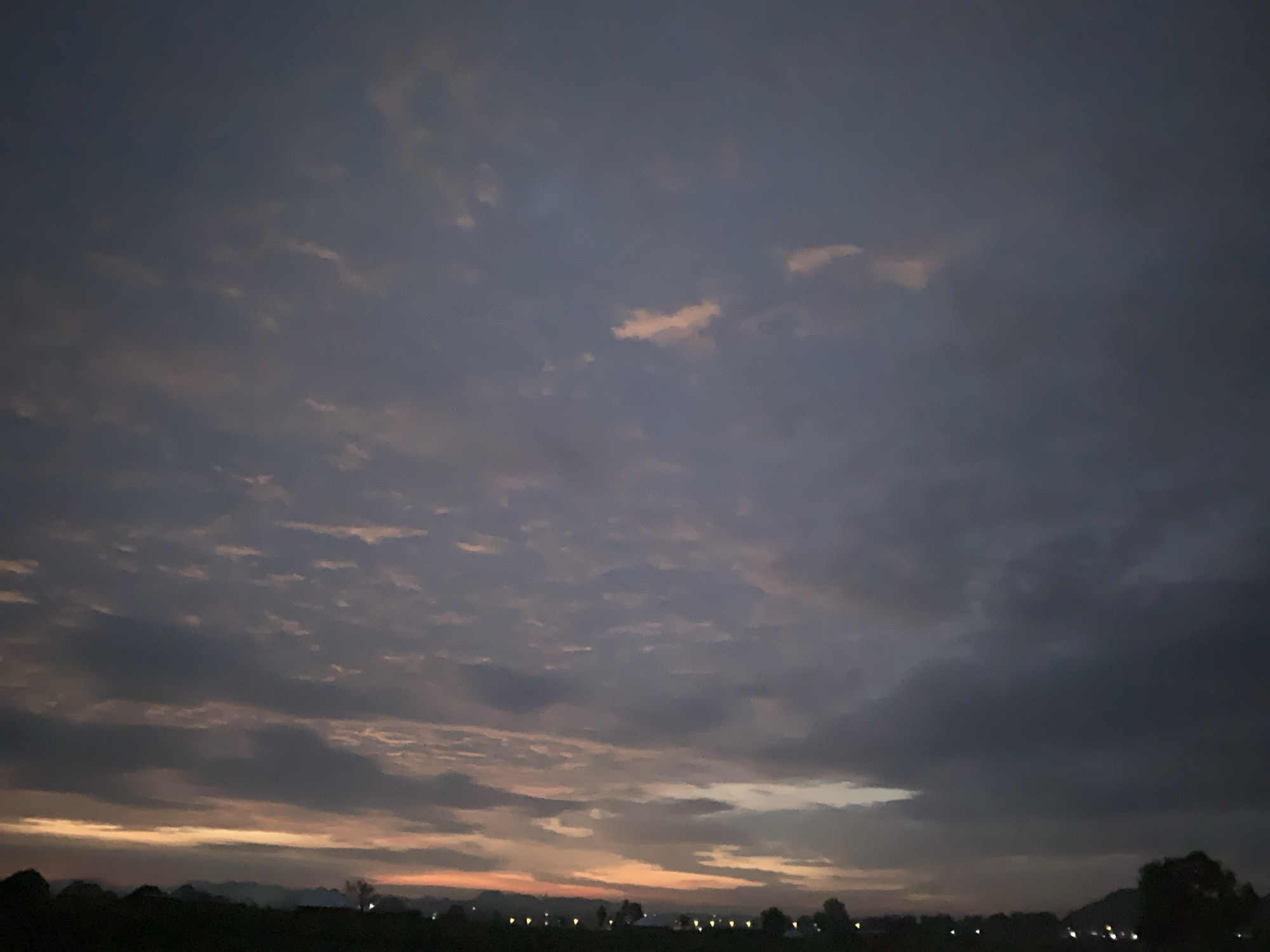 The image depicts a dusk sky filled with a mix of dark grey and lighter, pinkish clouds. The horizon line is low, showing a dark silhouette of trees and a few scattered lights, possibly from a town or village. The lighting suggests the sun has recently set, casting a warm glow near the horizon. The overall scene is one of a rural or suburban landscape at twilight in Kuru Karama, Nigeria.