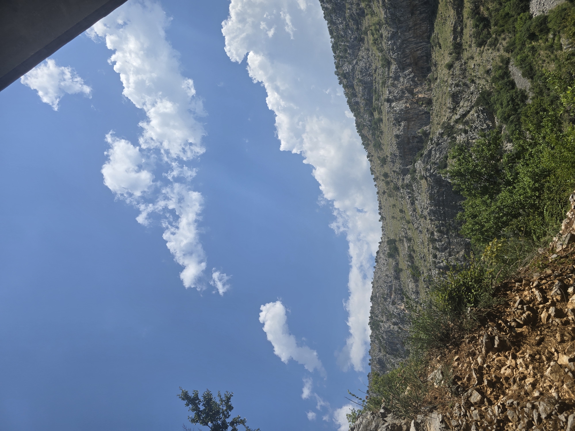 The image depicts a mountainous landscape under a bright blue sky with scattered white clouds. In the foreground, a steep, rocky slope is visible, covered with sparse vegetation, including bushes and small trees. Further back, the terrain rises into a larger, rugged mountain with sheer cliffs and rocky outcrops. Patches of green vegetation, including trees, are scattered across the mountain slopes. The overall environment suggests a natural, undeveloped area. The image does not contain any people, specific urban structures, or identifiable landmarks that would allow for precise localization within Podgorica, Montenegro. The perspective suggests a view from a lower elevation looking upwards towards the mountains.