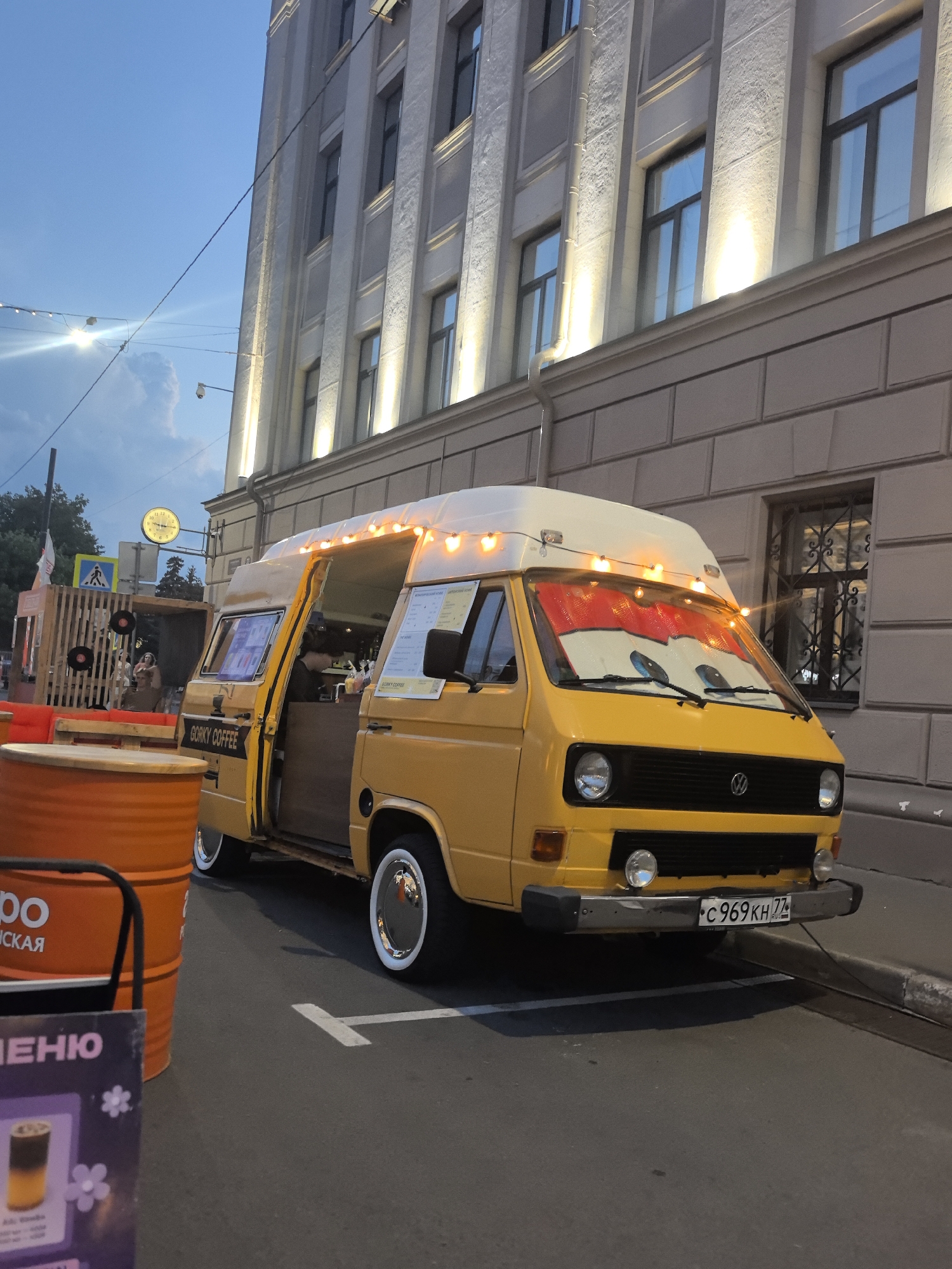 A yellow Volkswagen van, repurposed as a mobile coffee shop named "Gorky Coffee," is parked on a street in Nizhny Novgorod, Russia. The van has its side door open, revealing a counter and a person working inside. Decorative lights are strung across the top of the van, and a sunshade with cartoon eyes is placed on the windshield. In the foreground, an orange metal barrel and a menu board are visible. The background features a building with multiple windows, illuminated from below. The sky is a deep blue, suggesting it's either early morning or evening.