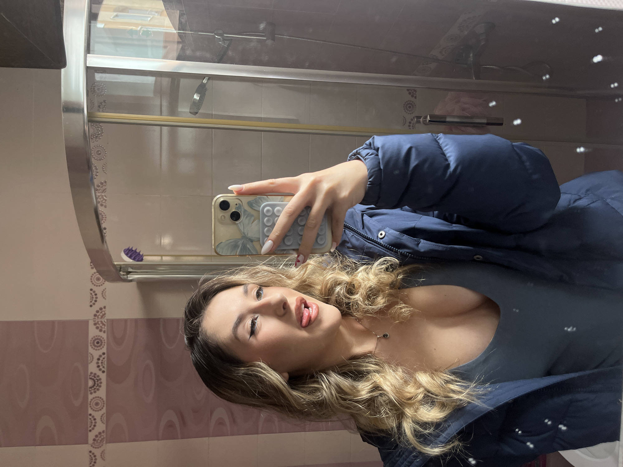 A young woman is taking a selfie in a bathroom. She is wearing a blue puffer jacket and a grey top. She has blonde curly hair and is sticking her tongue out. The bathroom has pink and white patterned tiles and a shower with a glass door. The location is likely within a residence and there is not enough context to determine if the location is in Sofia, Bulgaria.