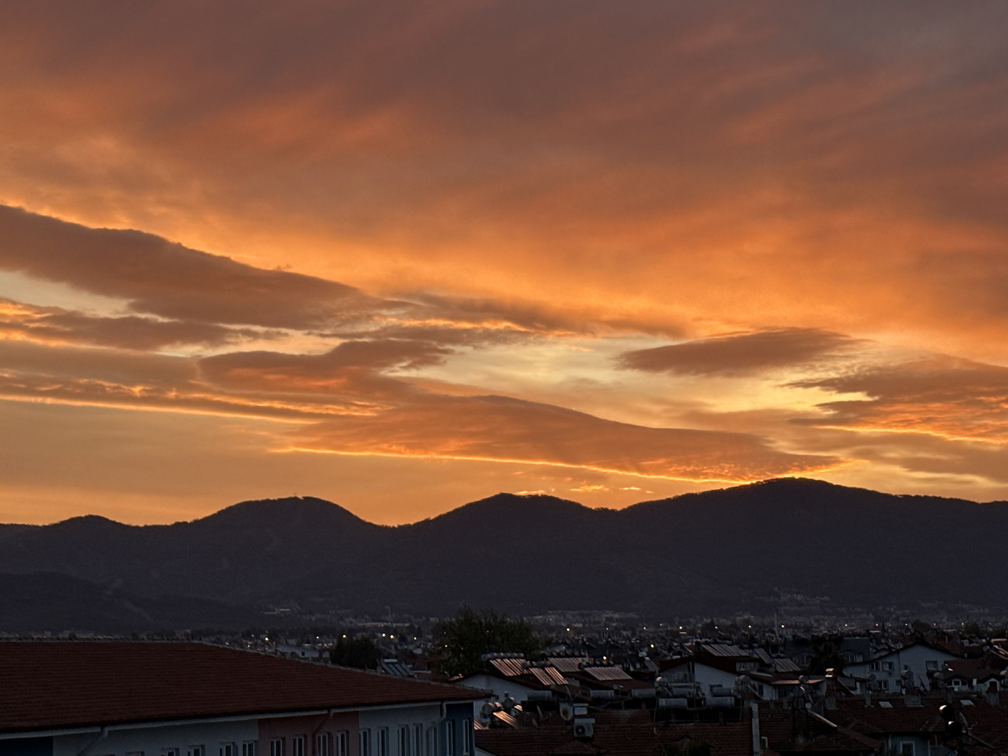 The image captures a dramatic sunset over Fethiye, Turkey. The sky is filled with orange and pink-hued clouds. Below the sky, a dark silhouette of mountains stretches across the horizon. The cityscape of Fethiye is visible at the base of the mountains, with rooftops of houses creating a dense pattern. In the foreground, part of a building with a red tiled roof and white walls is visible. The scene lacks people and focuses on the natural beauty of the sunset and the town's landscape.