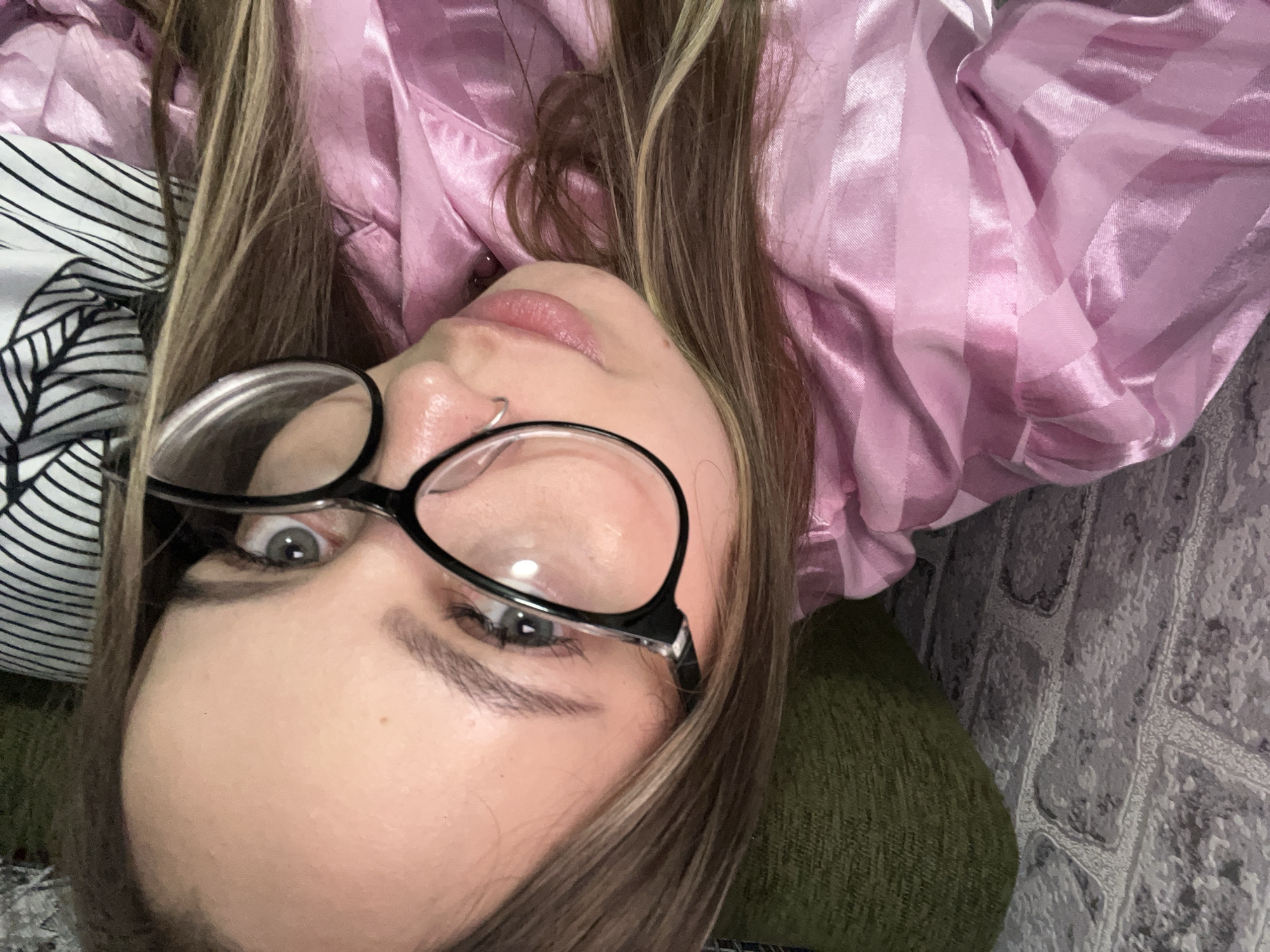 A woman with light brown hair and gray eyes is lying down, wearing black glasses and a pink striped shirt. She has a nose ring. She is lying on a green and white patterned surface, with a gray brick-like surface visible in the background. The photo is taken at a tilted angle, close to the woman's face. There is no indication of this image being in Novopokrovskaya, Russia.